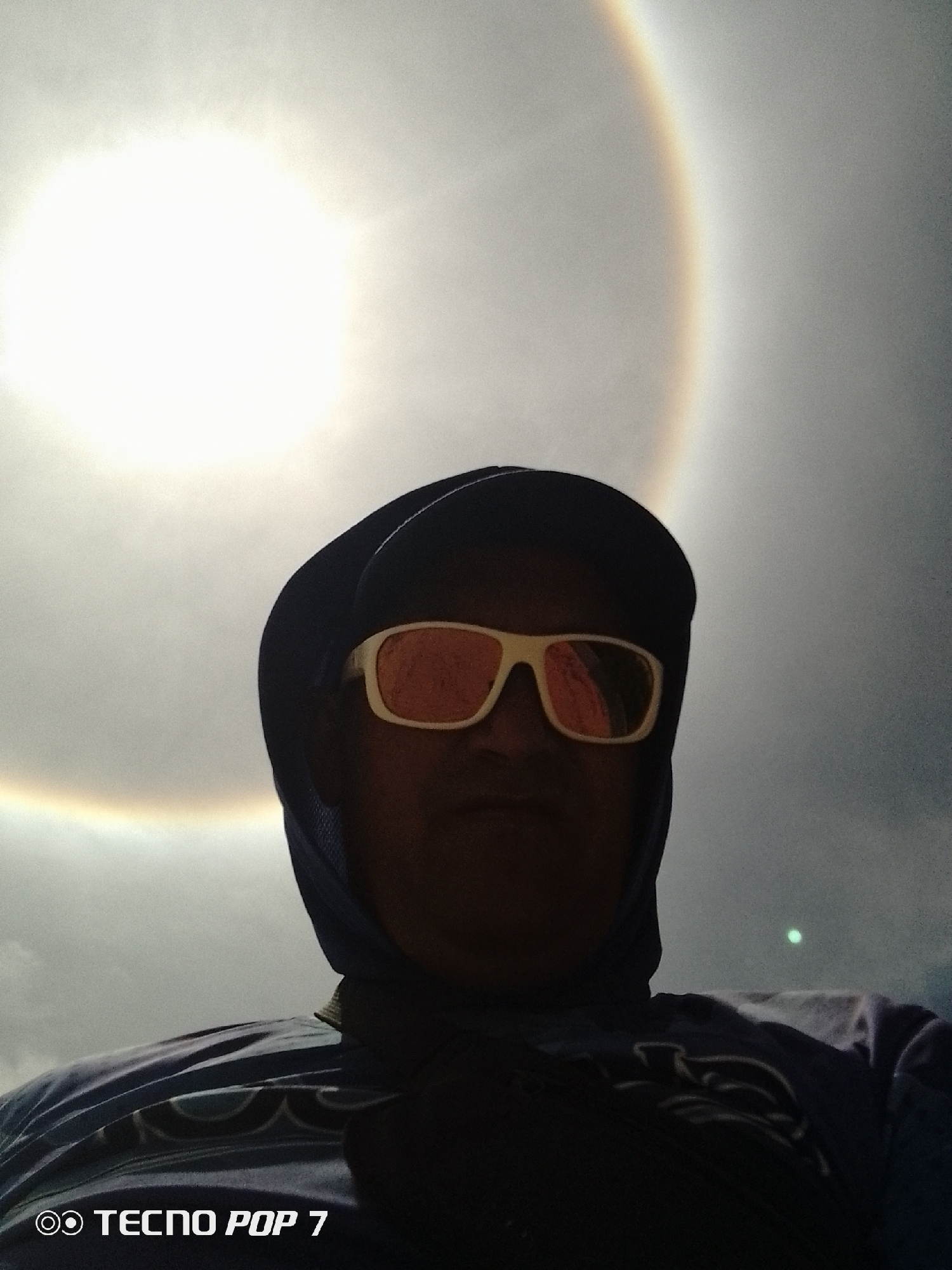 A person is wearing sunglasses and a hooded shirt in an outdoor setting in Pedro González, Venezuela. A halo is visible around the sun in the sky. The person is facing the camera.
