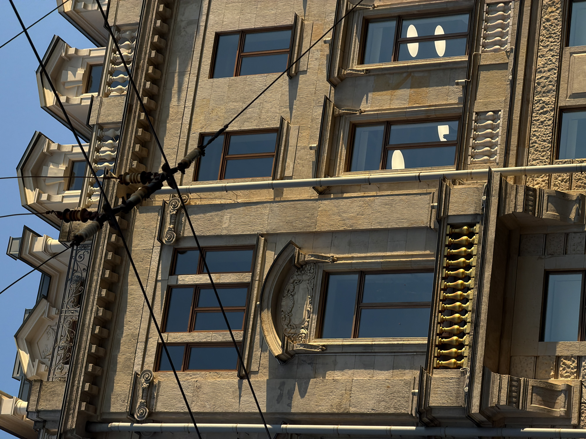 This is a close-up, eye-level exterior shot of a stone building in Saint Petersburg, Russia, bathed in the warm, golden light of late afternoon or early morning. The building features ornate architectural details, including layered cornices, decorative carvings above windows, and a balcony with a yellow balustrade. Several dark overhead wires, likely for public transport, crisscross the frame, casting shadows on the facade and obscuring parts of the upper floors. The sky above is a clear, bright blue. There are no people or vehicles visible in the image. The scene captures a typical urban streetscape in Saint Petersburg, characterized by its historic architecture and the ubiquitous presence of overhead utility lines.