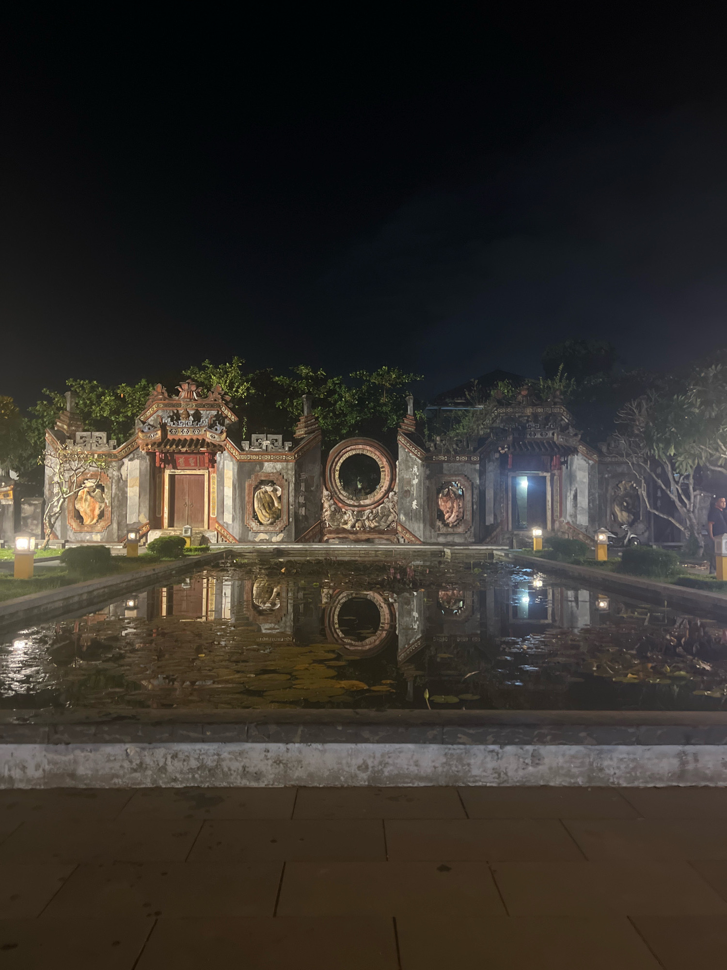 This is a nighttime, full-frame, eye-level shot of the exterior of the Temple of Literature in Hoi An, Vietnam. The temple is illuminated by warm, yellow lights, highlighting its intricate architecture. A still pond with lily pads occupies the foreground, reflecting the temple's facade and the night sky. The temple itself features ornate carvings and statues, with a central doorway leading into darkness. Trees surround the temple, adding to the serene atmosphere. The ground in front of the pond is paved with square tiles. A few people are visible in the background, standing near the temple entrance and to the right of the frame.