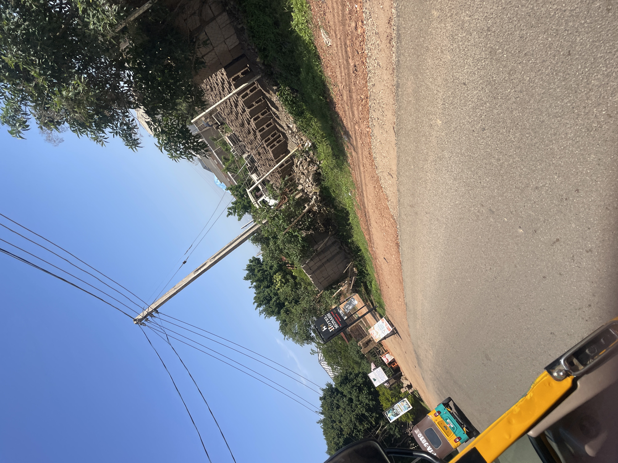 The image captures a street scene in Gura Top, Nigeria, under a clear blue sky. The perspective is from within a yellow and brown auto-rickshaw, showing a portion of its frame in the foreground. The road is asphalt and appears to be in moderate condition, with a dirt shoulder on the right side of the image.
Along the roadside, there is a construction site with stacks of concrete blocks. Several signs are visible. One prominent sign reads "HOTEL MONASH". Other smaller signs are also present, some with graphics, suggesting commercial activity.
Mature trees with dense green foliage line the area behind the construction site. Power lines are strung across the sky, supported by a wooden utility pole. In the distance, more trees and buildings are partially visible, indicating an urban or semi-urban setting. A teal and yellow auto-rickshaw is traveling ahead on the road, with its license plate partially visible. The overall impression is of a sunny day in a developing area.