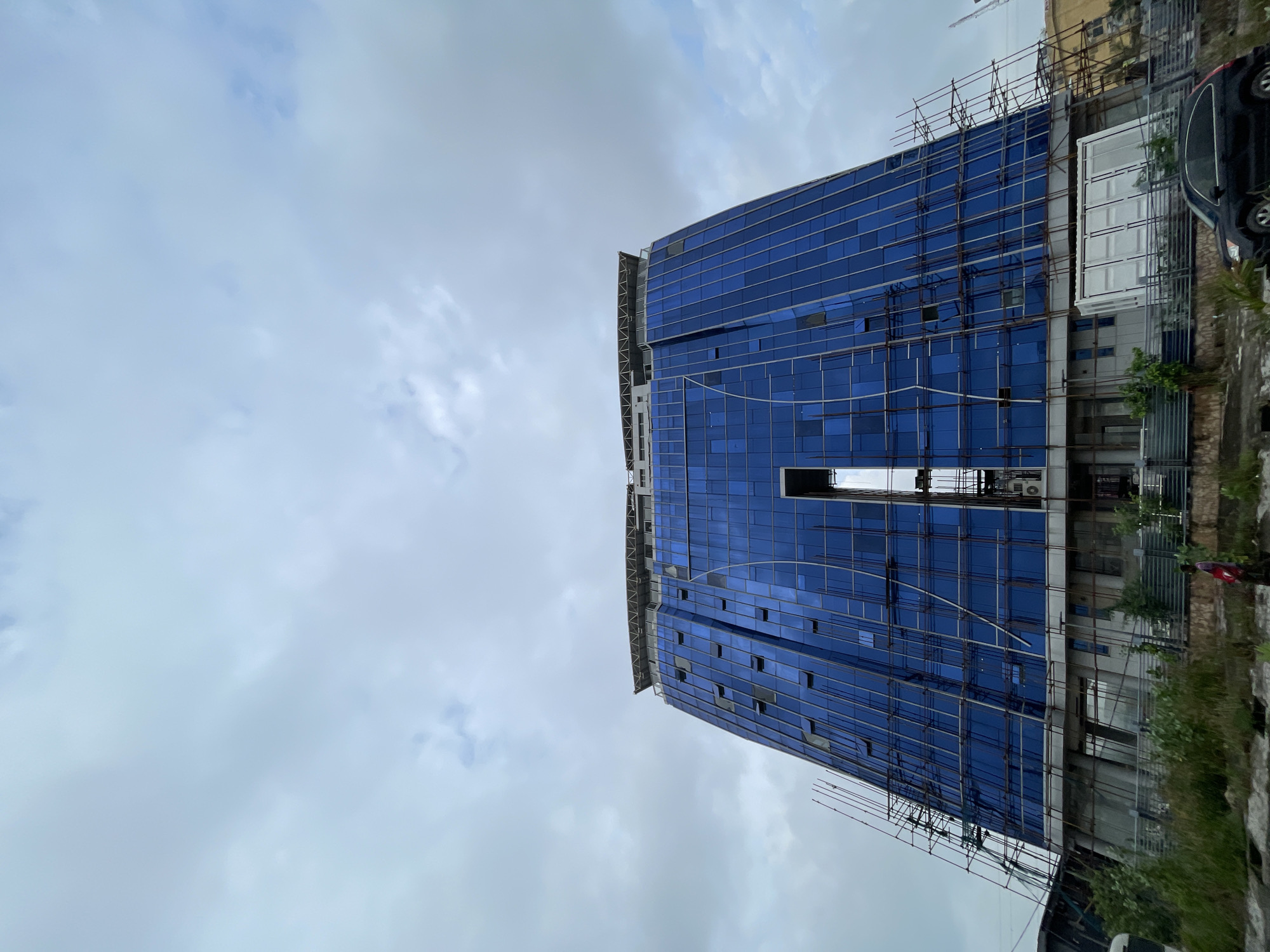 A low-angle, full shot captures a large, blue glass-paneled building under construction in Ilado, Nigeria, against a cloudy sky. The building has a distinctive, curved, trapezoidal shape, wider at the bottom and narrowing towards the top, with a central vertical cutout revealing scaffolding. Scaffolding structures are visible on both sides of the building, suggesting ongoing work. Multiple windows on the facade are open. At the very top, a flat roof structure with a crisscross pattern is present, potentially a helipad or rooftop amenity. In the foreground, a patch of overgrown green vegetation and some dirt paths are visible. A person in a pink shirt and dark pants walks on a path to the left of the frame, and a black SUV is parked on the right side. The overall environment appears to be an urban or semi-urban setting undergoing development.
