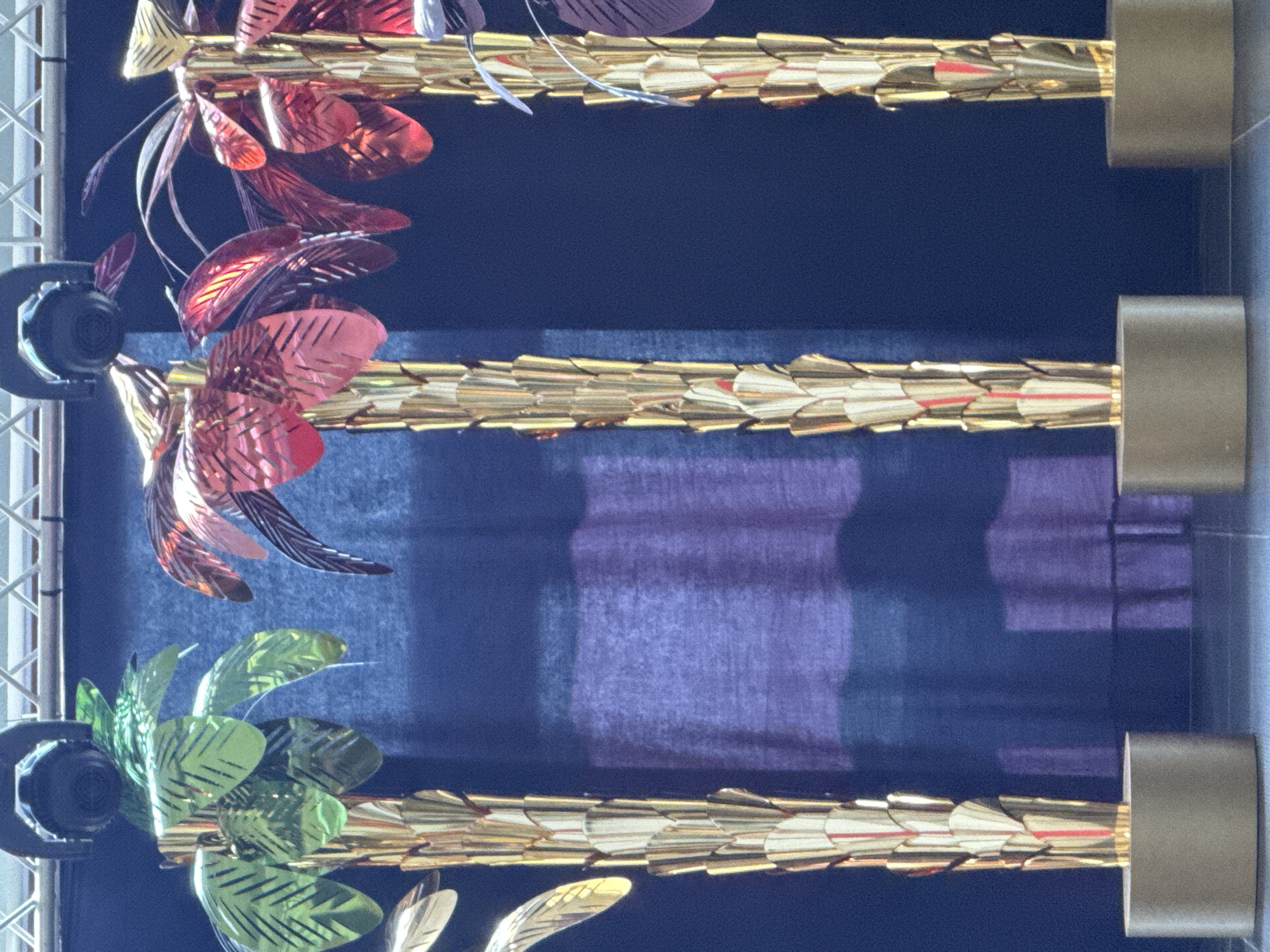 This media file displays a close-up view of decorative elements, likely part of an artistic installation or architectural feature. The subjects are primarily composed of metallic, leaf-like or feather-like ornaments in shades of red and green, attached to golden, segmented rods. These rods are supported by cylindrical metallic fixtures, one of which is partially visible on the left side of the image, appearing to be a spotlight or a mounting bracket. The background is a dark, textured fabric, possibly a curtain or a wall covering, with horizontal bands of lighter purple and blue hues, suggesting light filtering through or reflections.
There is no activity or event taking place; the image is a still capture of static decorative objects. Notable details include the shimmering, reflective quality of the metallic decorations, highlighting their artificial nature and intricate design. The lighting appears to be artificial, casting shadows and illuminating the metallic surfaces. The overall impression is one of ornate decoration, possibly for an indoor event or a theatrical setting.
There is no visible text in the image. The location context of Sochi, Russia, might suggest this installation is part of a public space, a hotel, or a venue within the city, but the image itself does not contain explicit cues to confirm this. The emotions evoked are neutral, focusing on the visual aesthetic of the decorative elements.