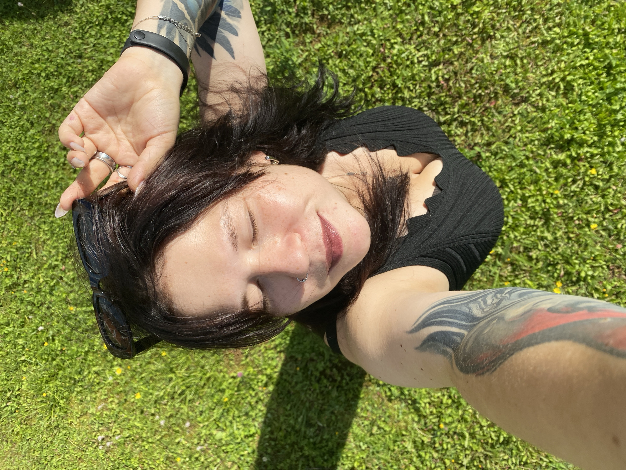 A high-angle, medium shot captures a young woman lying on lush green grass with small yellow flowers, possibly in the city of Фоки, Russia. Her eyes are closed, and her head is tilted back, with her right hand touching her hair. She wears a black, ribbed top with a scalloped neckline and has a nose ring. Tattoos are visible on her left arm and right arm. She also wears rings on her right hand and a black wristband. Sunglasses are perched on her head. The sun casts a shadow on the grass to her left.