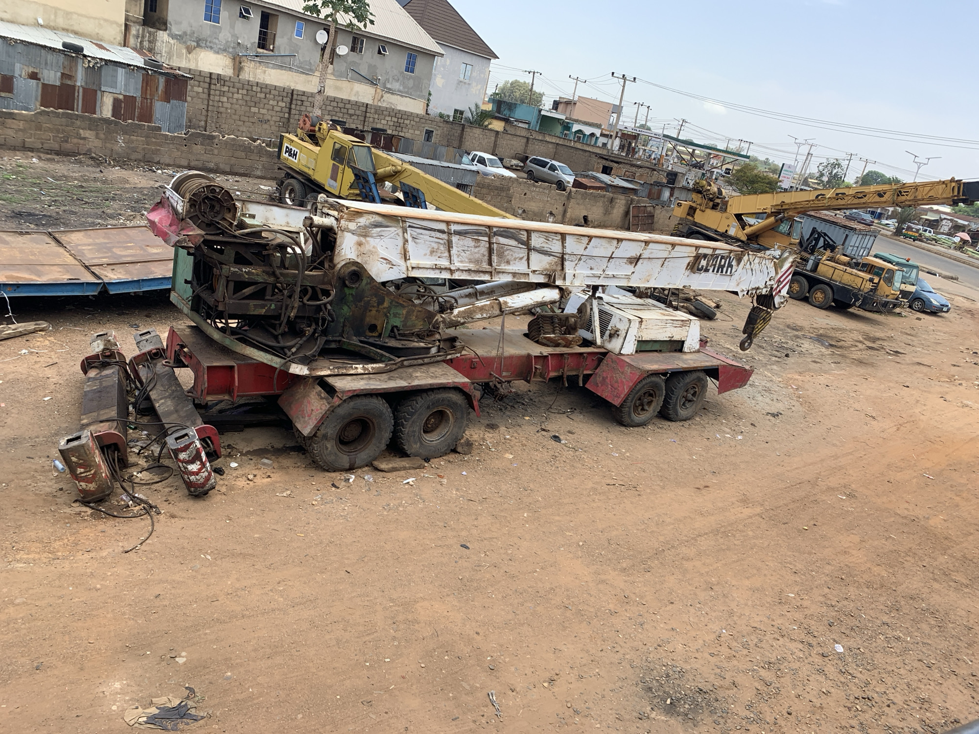 The image depicts a construction site in Jos, Nigeria, showcasing heavy machinery. A dilapidated red and white crane with rusted parts is in the foreground. Nearby are disconnected outriggers. In the background, there are other pieces of construction equipment, including a yellow P&H crane and a yellow crane truck, parked near a building. A brick wall runs along the top of the frame, with various buildings and vehicles visible beyond it. The ground is dirt and gravel. The sky is overcast.