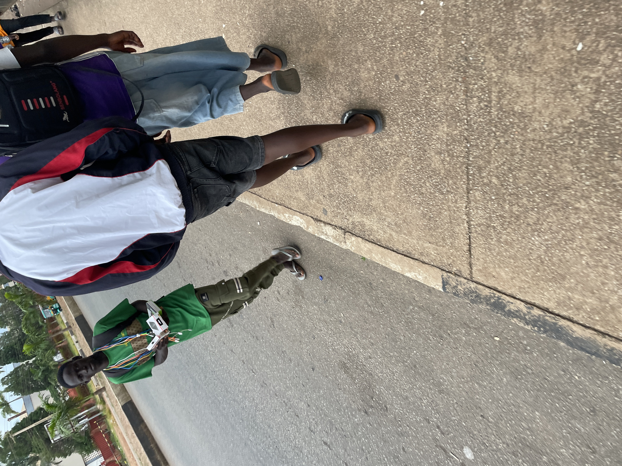 In Jos, Nigeria, a young man in a green t-shirt and olive cargo pants walks along the side of a road, holding a white box. He has a backpack on his shoulders and colorful lanyards around his neck. Two other individuals are walking ahead of him on the sidewalk, one wearing a purple shirt and light blue shorts, the other in a jacket and dark shorts. A three-wheeled vehicle is visible in the background, parked on the side of the road. The scene appears to be a typical street view in the city.