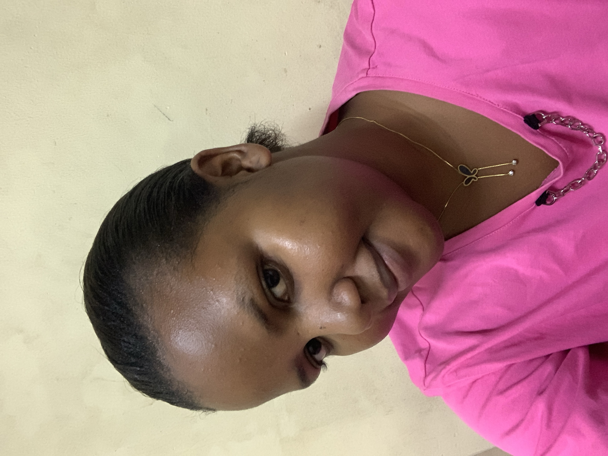 A close-up, tilted portrait captures a dark-skinned individual with sleeked-back black hair, wearing a bright pink top. A delicate gold chain with a butterfly pendant is visible around the person's neck. The backdrop is a plain, pale yellow wall. This image does not contain enough information to determine a specific location in Dar es-Salaam, Tanzania.