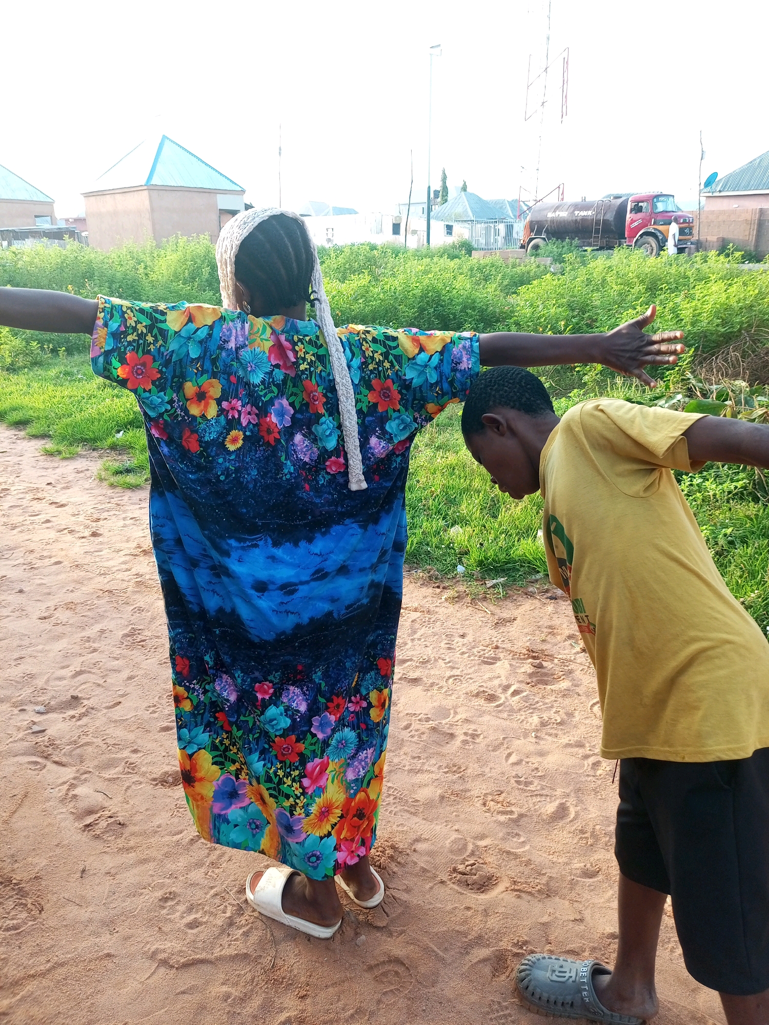 The image depicts two individuals standing on a sandy path in Tempure, Nigeria, with their arms outstretched. A person wearing a long, colorful floral-patterned dress and a white head covering stands with their back to the camera, arms spread wide. To their right, a person in a yellow t-shirt and black shorts bends forward, also with arms extended. The environment includes green vegetation, sandy ground, and buildings in the background. A red and silver water tank truck is visible further in the background, alongside other structures. The sky is bright, suggesting daylight.