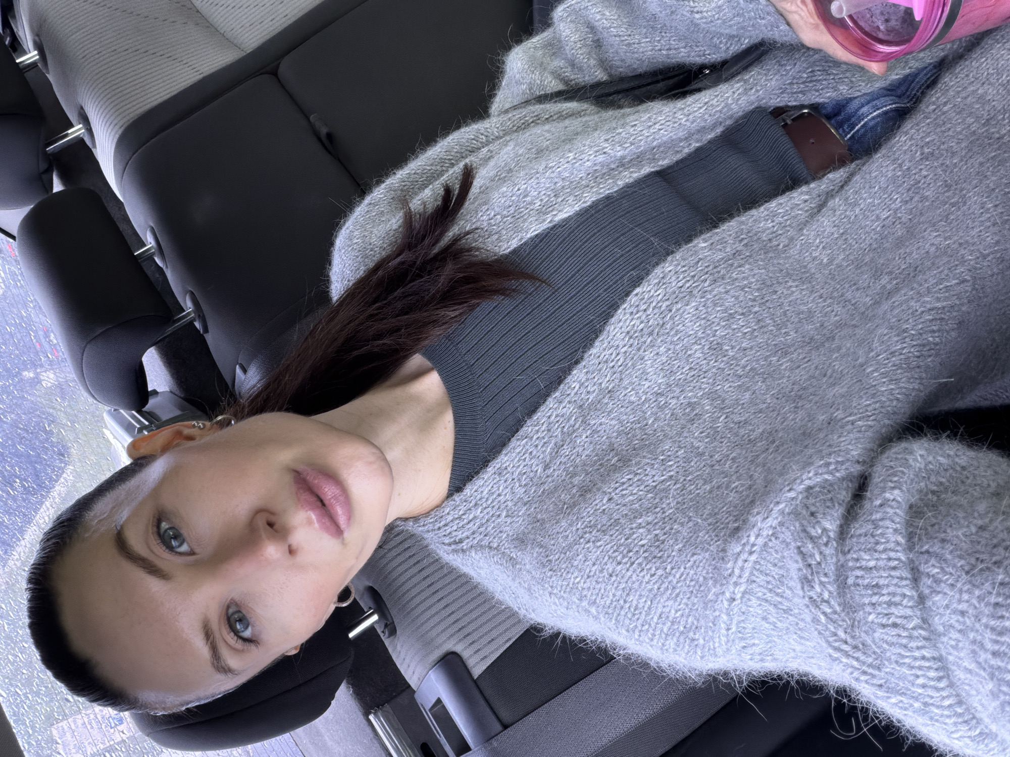 A woman is sitting in the back seat of a car. She has dark hair and blue eyes, wearing a gray sweater. She appears to be holding a pink water bottle. The seat is gray and black. The image does not provide information to identify the location as Perm, Russia.