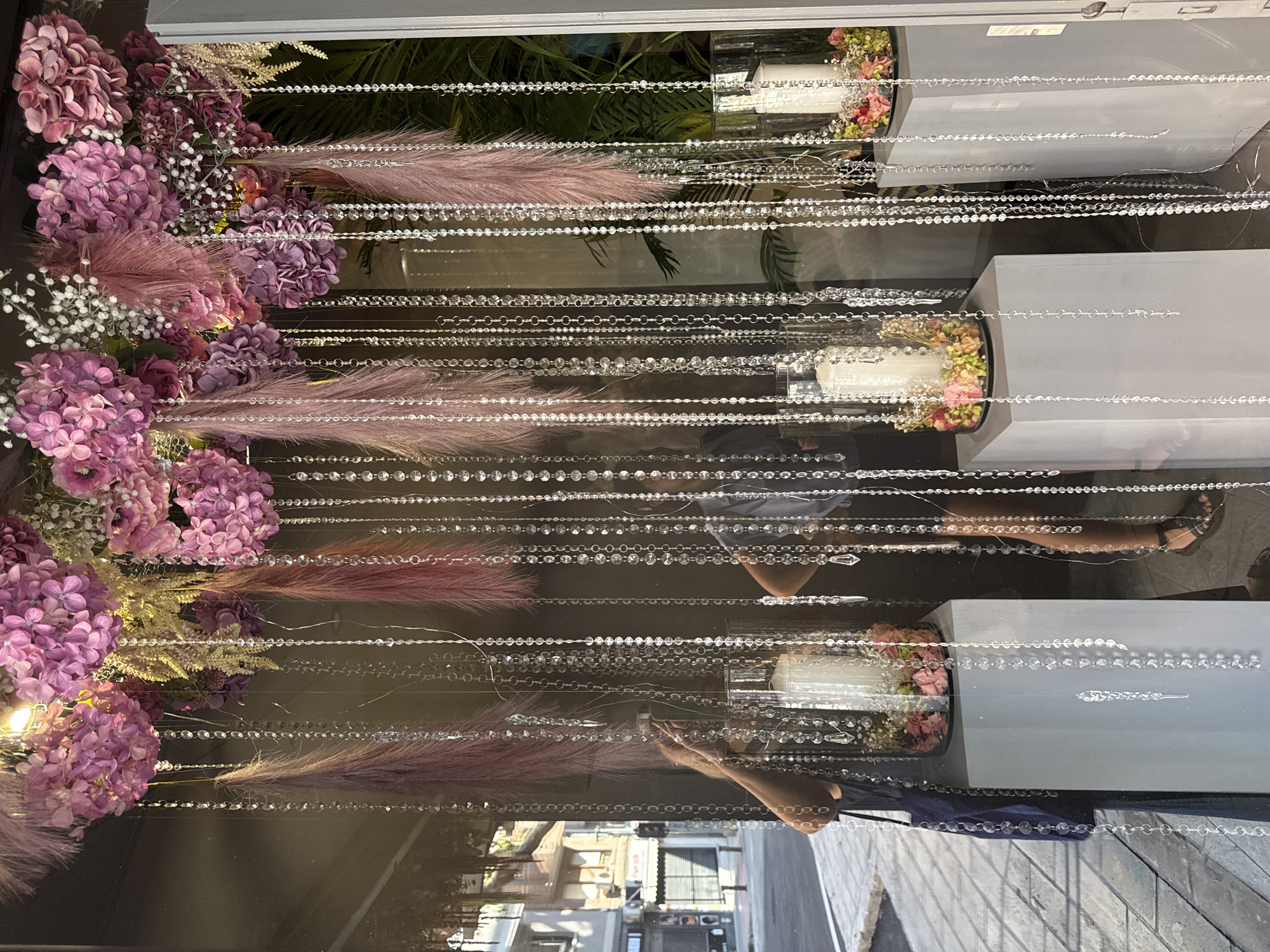 The image displays an elegant decorative arrangement, likely viewed through a reflective glass panel or window, as indicated by visible reflections. On the left side of the frame, lush clusters of purple and mauve hydrangeas are prominent, intermingled with delicate white baby's breath and soft, feathery plumes of pinkish-purple, possibly pampas grass. These floral and botanical elements appear to be arranged vertically along the left edge.
Numerous strands of sparkling clear beads, resembling crystal chains, stretch horizontally across the frame. These beads vary in style, with some being continuous strings of small spherical beads, while others feature larger, faceted crystals or segments of silver-toned chain links. They create a shimmering, curtain-like effect that partitions the display.
Behind this beaded screen and floral decor, several plain grey, rectangular stands are positioned vertically along the right side of the image. At least two of these stands support cylindrical glass vases, each containing a mixed floral arrangement of pink and white flowers, centered around a white candle.
Notable details include reflections on the glass surface. Partial views of an outdoor urban environment are visible, showing buildings and streets, suggesting a daytime setting. Reflections of people are also apparent, including a person's arms and hands near the bottom center, and another person's lower legs and feet wearing sandals towards the bottom right. The green foliage of what appear to be palm leaves is also reflected, either from the outdoor environment or as part of the interior decor behind the glass. No discernible text is visible in the image. The overall impression is one of sophisticated decor, potentially a storefront display or an elaborate interior design.