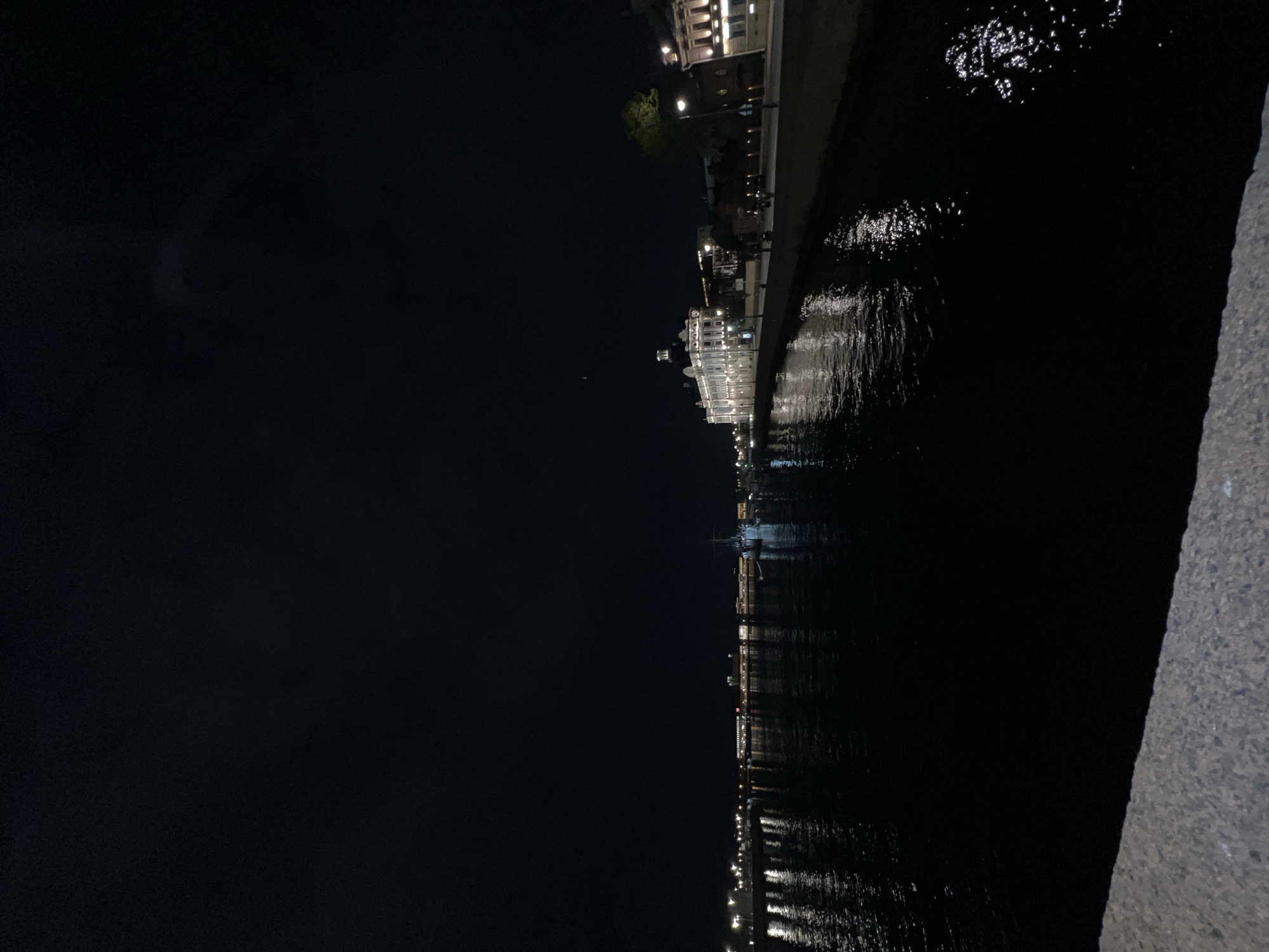 Captured at night, the image depicts a waterfront scene in Saint Petersburg, Russia. The cityscape, illuminated by artificial lights, stretches along the water's edge. A body of water reflects the city lights, creating streaks of brightness on the dark surface. To the left, a dark expanse suggests the night sky, with a faint crescent-shaped light feature. The right side of the image features a textured surface, possibly a stone embankment or wall.
