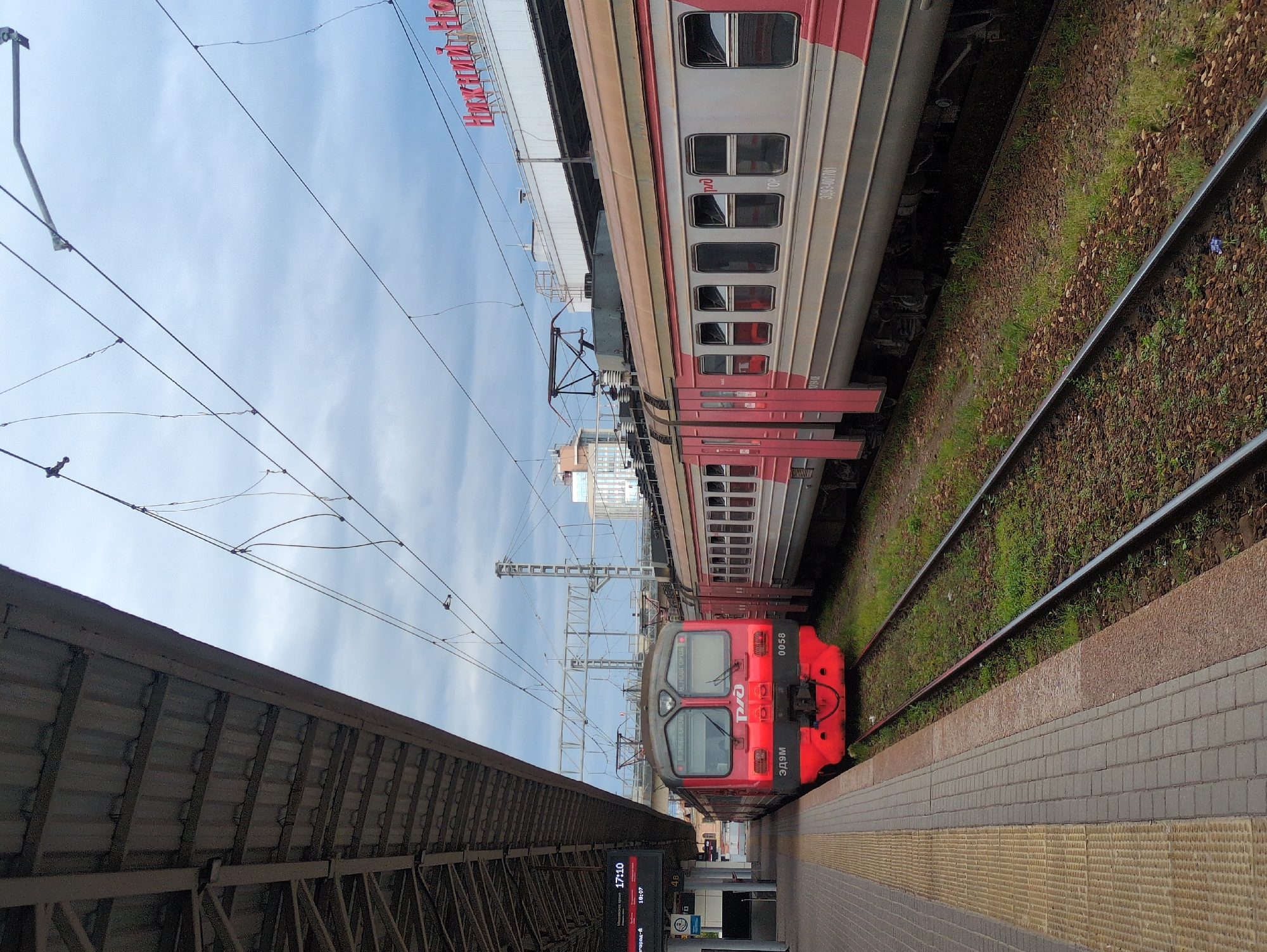 The image depicts a train station platform in Nizhny Novgorod, Russia, under an overcast sky. Two trains are stopped on adjacent tracks. The train on the left is a red electric train with the Russian Railways logo visible on its front. The train on the right is a passenger train with red and gray carriages. Overhead power lines stretch across the scene. In the background, a building with red signage spelling out "Нижний Новгород" (Nizhny Novgorod) is visible, confirming the location. A digital display board on the platform shows departure times. The platform itself is paved with brick and features a yellow tactile warning strip along the edge. The tracks are lined with ballast and overgrown with grass.