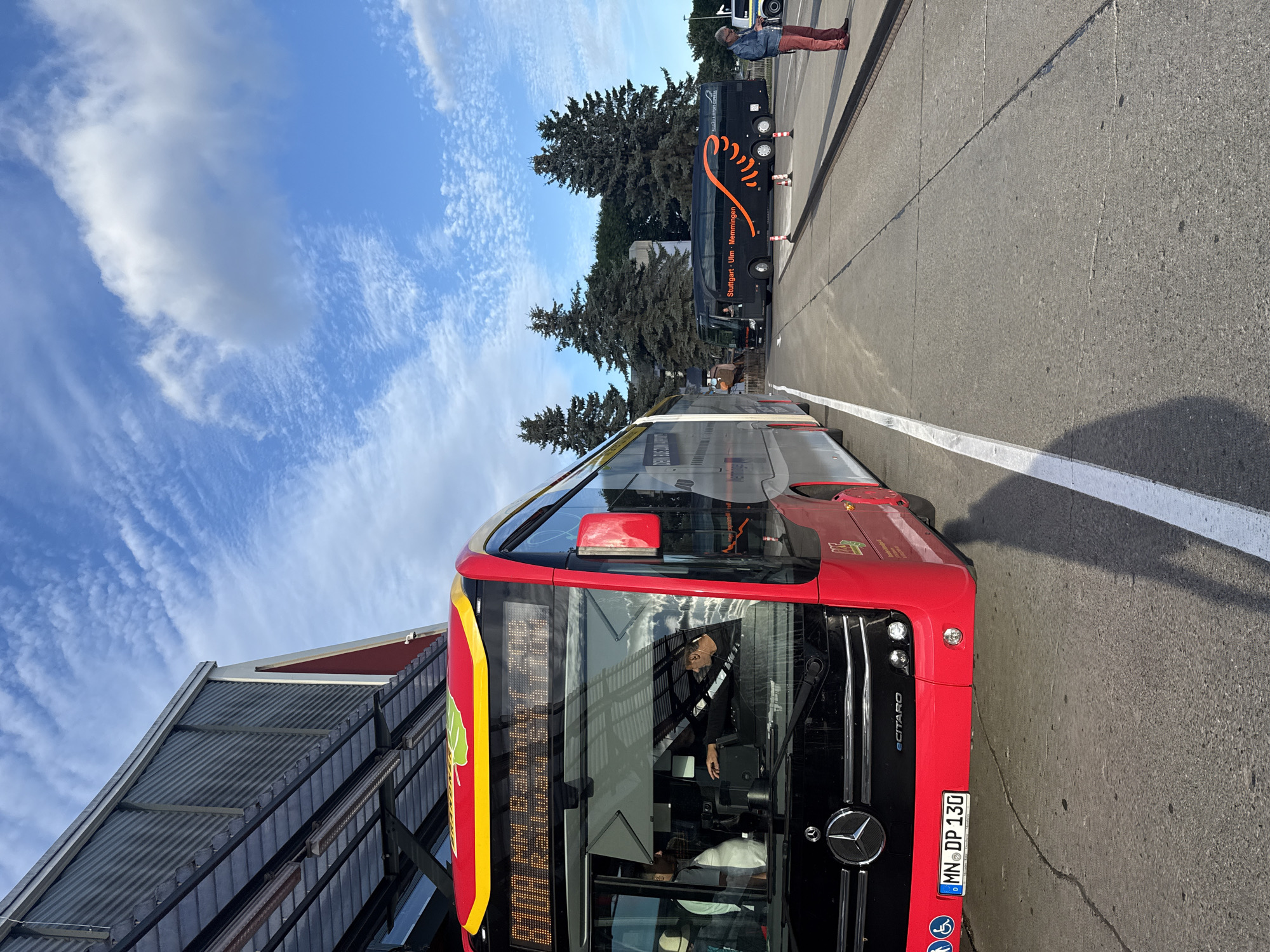 This image was taken at the bus station in Künersberg, Germany. In the foreground, a red Mercedes-Benz eCitaro bus is parked. The bus has a digital display showing "München Hbf Bahnhof". A license plate reading "MN DP 130" is visible on the front of the bus. The driver, a man wearing a dark shirt, is seated inside the bus, seemingly operating the vehicle. In the background, a black coach bus with orange accents is parked. This coach displays "Stuttgart - Ulm - Memmingen" and "ALLGÄU AIRPORT EXPRESS". A person in red pants and a blue jacket stands near the coach bus. The scene is set against a backdrop of a bright blue sky with scattered clouds and several tall evergreen trees. The environment appears to be an open-air bus terminal with asphalt ground and a building structure partially visible on the left.