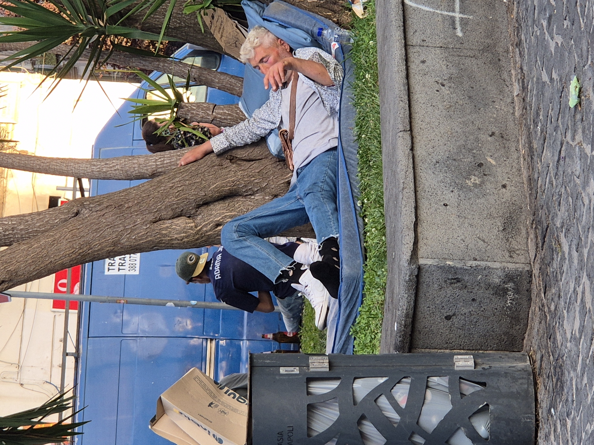 In Naples, Italy, a man with light-colored hair is lying on his back on a blue mat, which is placed on the grass. He is wearing a patterned shirt, blue jeans, and white sneakers with black socks. His right arm is raised, and he appears to be holding something near his face. A bottle of water and a newspaper or book are on the mat next to him. In the background, a blue van is parked, and two individuals are visible. One person, wearing a dark shirt and a green cap, is sitting down. Another person, partially obscured by a tree, appears to be a child with dark hair. A trash can with an open lid is situated to the left of the scene. The environment includes trees, grass, and a stone bench. The ground is paved with cobblestones.