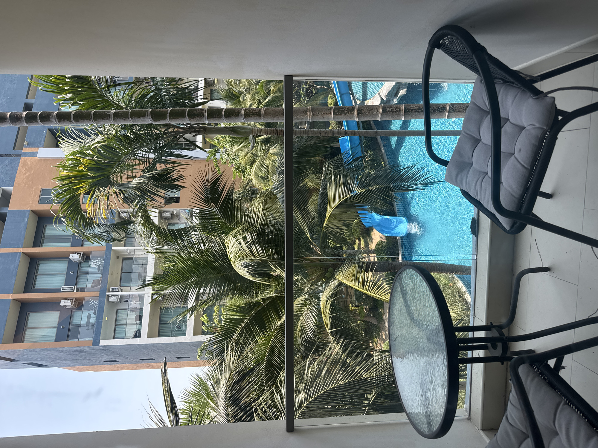 The image depicts a scene from a balcony in Pattaya City, Thailand. The view overlooks a swimming pool area with blue water, a slide, and various palm trees that are obstructing some of the surrounding buildings. On the balcony itself, there is a glass-topped table and two chairs with gray cushions. The architectural style of the adjacent building is modern, featuring colors such as blue, brown, and gray. The sky is visible, although the overall light suggests a bright, sunny day.