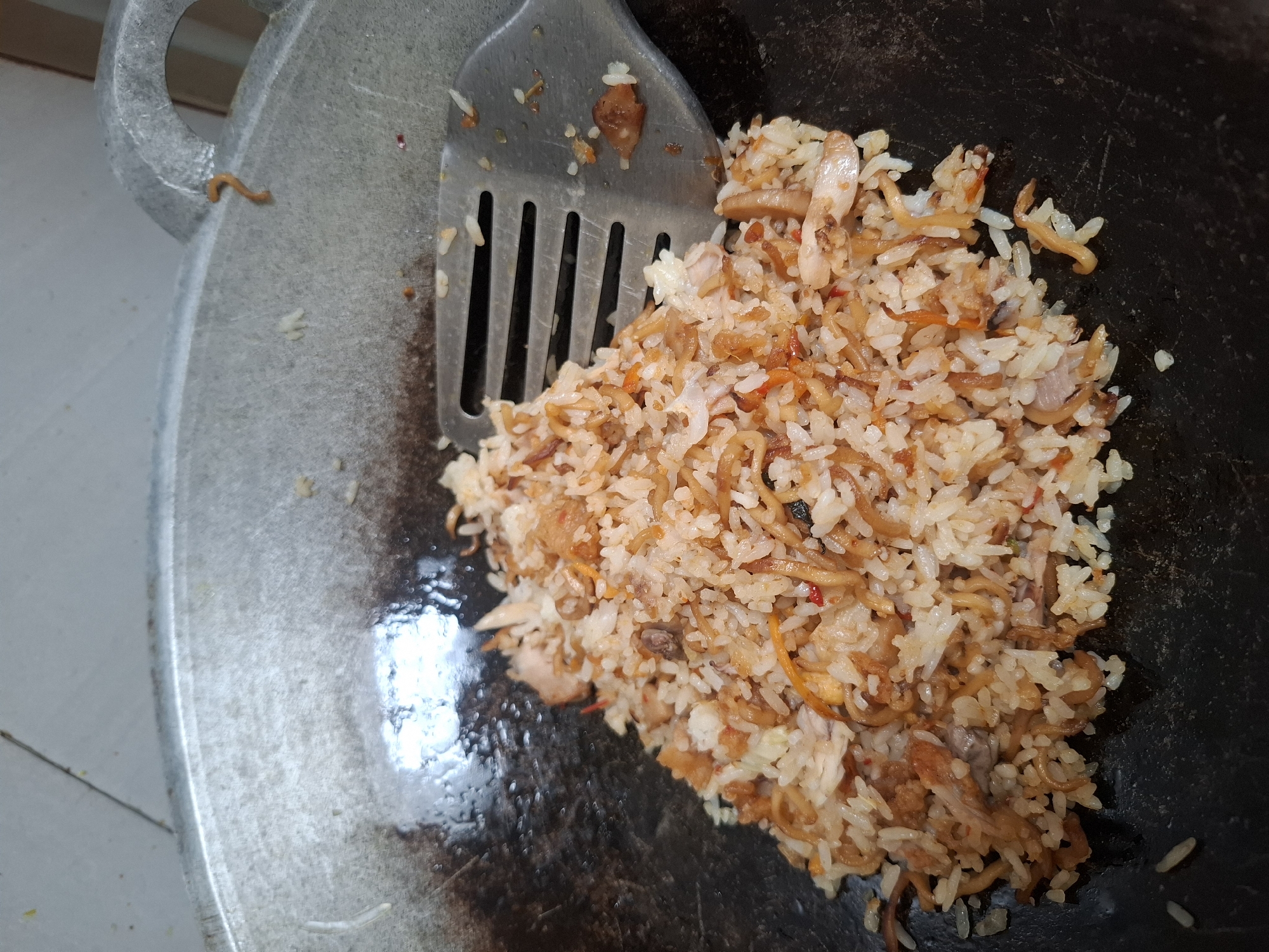 A close-up, overhead shot depicts a pile of fried rice in a dark, seasoned wok. The rice is mixed with small pieces of meat and vegetables, with some noodles intertwined. A metal spatula with slots is resting on the pile, partially obscuring it. The wok's rim is visible, showing signs of use and wear. Outside the wok, the tiled floor of what appears to be a kitchen is visible. The overall lighting is somewhat dim, creating a slightly grainy texture. The scene is likely from a home kitchen in Babakan Madang, Indonesia.