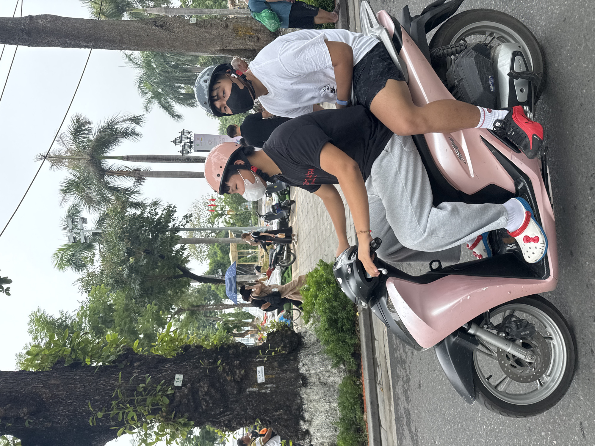 Two young Asian males are riding a pink scooter on a paved road in Hanoi, Vietnam. The rider in front is wearing a pink helmet, a white mask, and a black t-shirt with grey sweatpants. He has on white and blue slides with a red Hello Kitty character. The passenger behind him is wearing a grey helmet, a black mask, and a white t-shirt with black patterned shorts. He has on white socks and red and black sneakers. The scooter is a light pink color with black accents. In the background, there are palm trees, other scooters, and people walking. One person in the background is holding a blue umbrella. There is a large tree with a sign on it to the left of the frame. The sky is overcast.