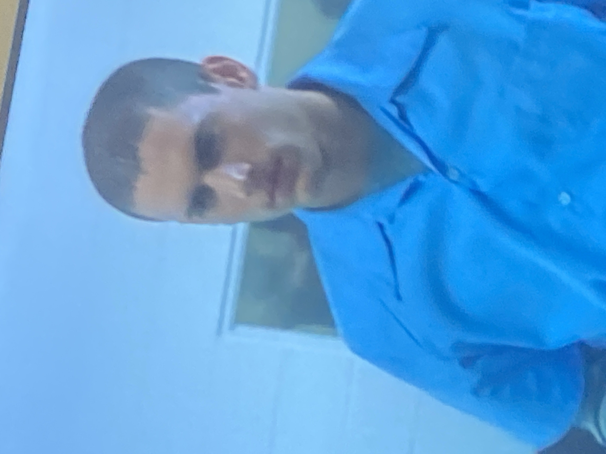 The image features a young man captured from the chest up, positioned against a light-colored, possibly indoor background that includes what appears to be a wall and a window or doorway. The man has very short, dark hair in a buzz-cut style and a medium complexion. He is wearing a light blue collared shirt. His expression is neutral, appearing somewhat serious or pensive, and he is looking slightly towards the viewer. The image is taken at a slight angle, with the man's head tilted. The lighting in the scene is gentle, casting subtle shadows on his face and clothing. There is no discernible specific activity taking place other than being photographed. The image quality is somewhat soft and grainy. No text is visible. While the location context is Gosa, Nigeria, no specific visual cues within the image confirm this.