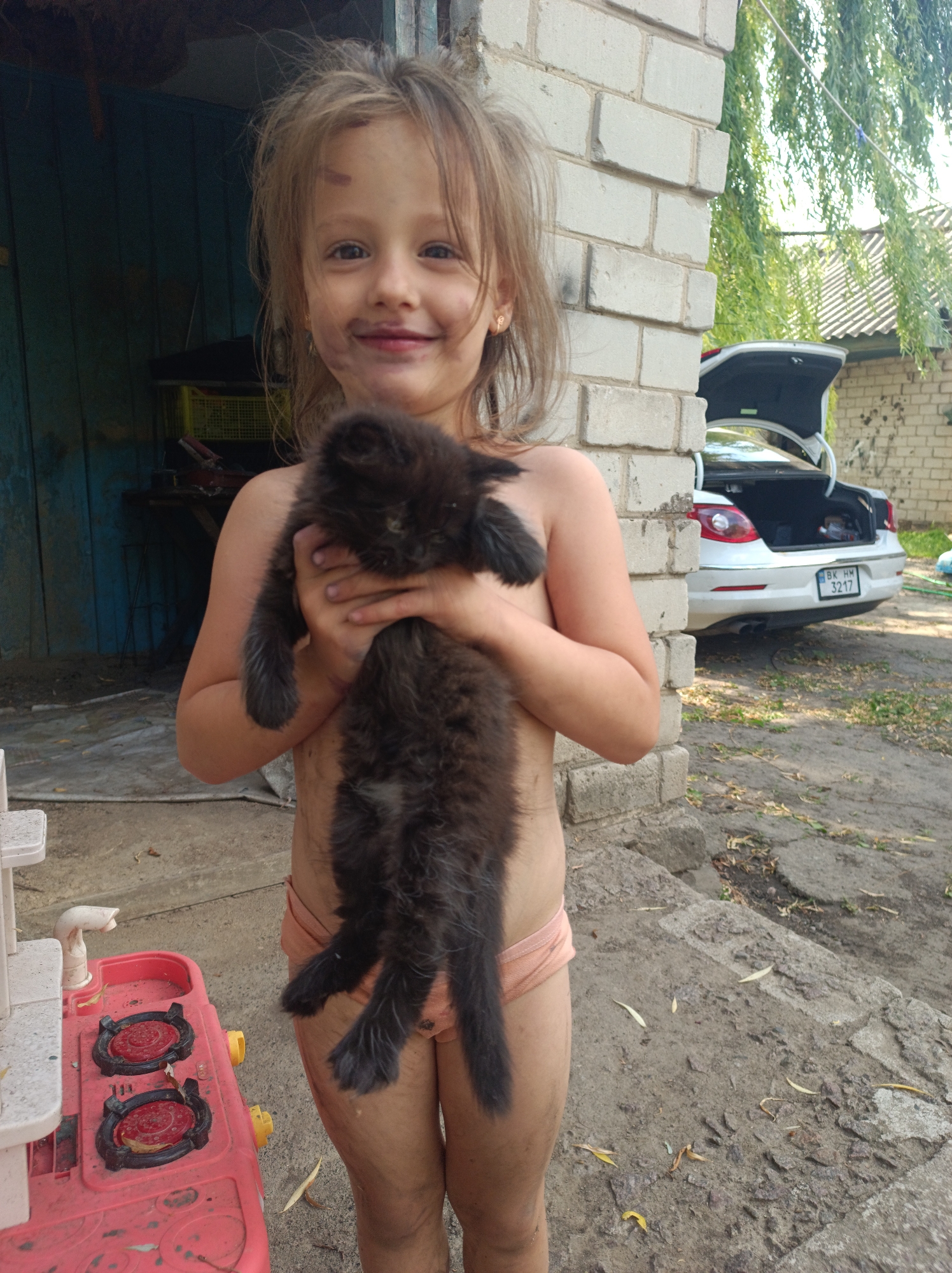 In Saksahan, Ukraine, a young girl with messy blonde hair, her face smudged with dirt, smiles at the camera while holding a fluffy black kitten. She is wearing light peach-colored underwear. The scene appears to be outdoors, possibly in a yard, with a brick building to her right. In the background, a white car with its trunk open is parked. To the girl's left, a red plastic toy kitchen set is visible. The ground is rough and appears to be a mix of dirt and concrete. The lighting suggests it is daytime, with sunlight filtering through the trees. The overall impression is a candid, outdoor moment capturing a child and her pet in a rustic setting.