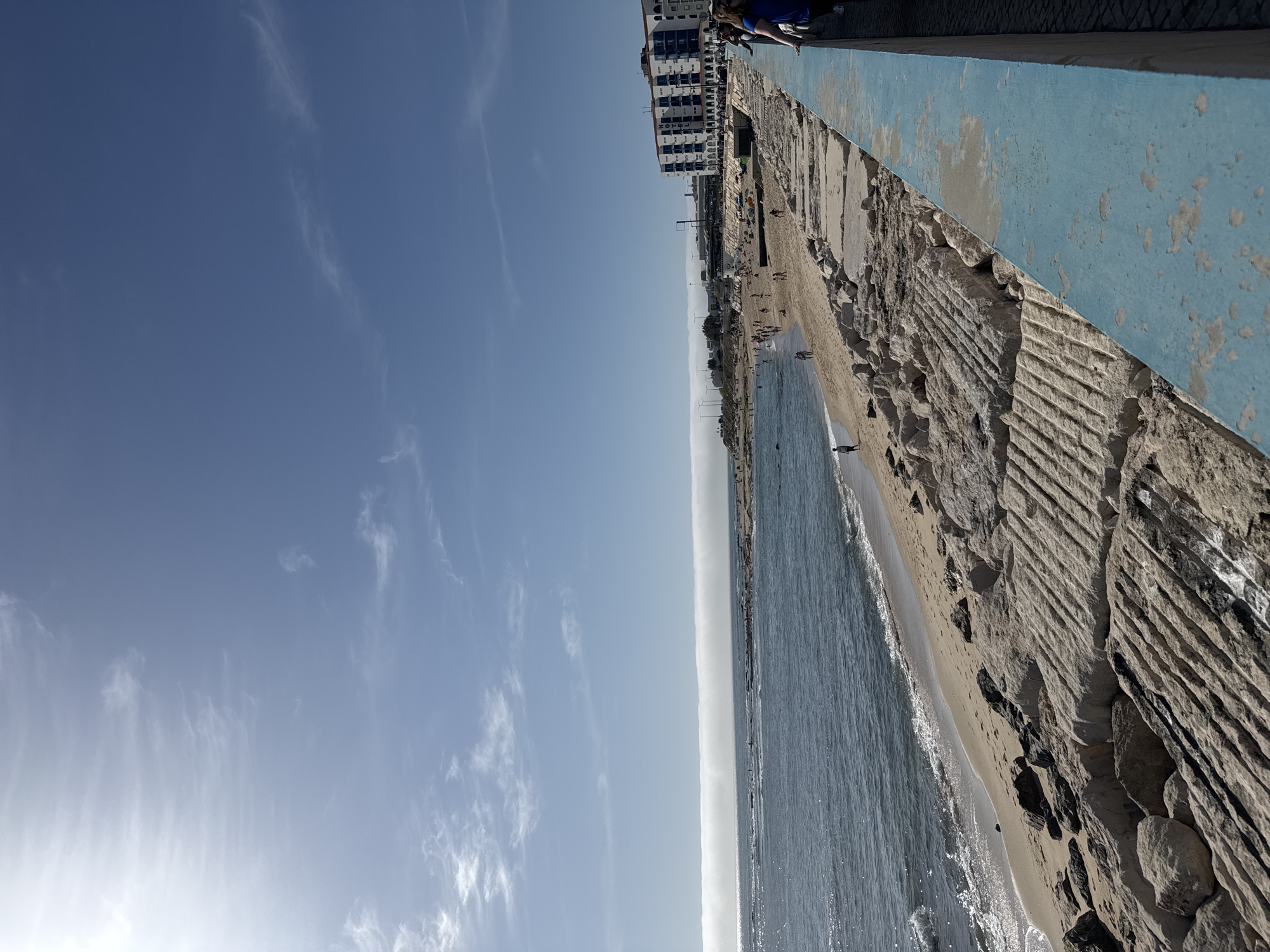 This image captures a vibrant coastal scene at Figueira da Foz, Portugal, on a bright, sunny day. The view is from a slightly elevated position, looking down towards a sandy beach, the ocean, and a bustling promenade.
In the foreground, a prominent concrete or rock seawall, featuring deep horizontal grooves, separates the beach from an elevated walkway. A section of this walkway's edge is painted light blue, with visible signs of weathering and peeling paint. A few individuals are seated or standing on this blue barrier.
The sandy beach stretches along the coastline, populated by several people enjoying the pleasant weather. Many individuals are scattered across the sand, some strolling closer to the water, while others are in the shallow, clear ocean where small waves are breaking.
Further along the coast, a line of multi-story buildings forms an urban backdrop, one of which clearly displays the text "HOTEL." In the distant background, faint structures resembling wind turbines are visible along the horizon. The sky is a vast expanse of clear blue, adorned with scattered, wispy white clouds. The sun, positioned towards the lower left, suggests it is mid-morning or early afternoon. The scene conveys a relaxed and lively atmosphere, with people engaging in leisurely activities by the sea.
