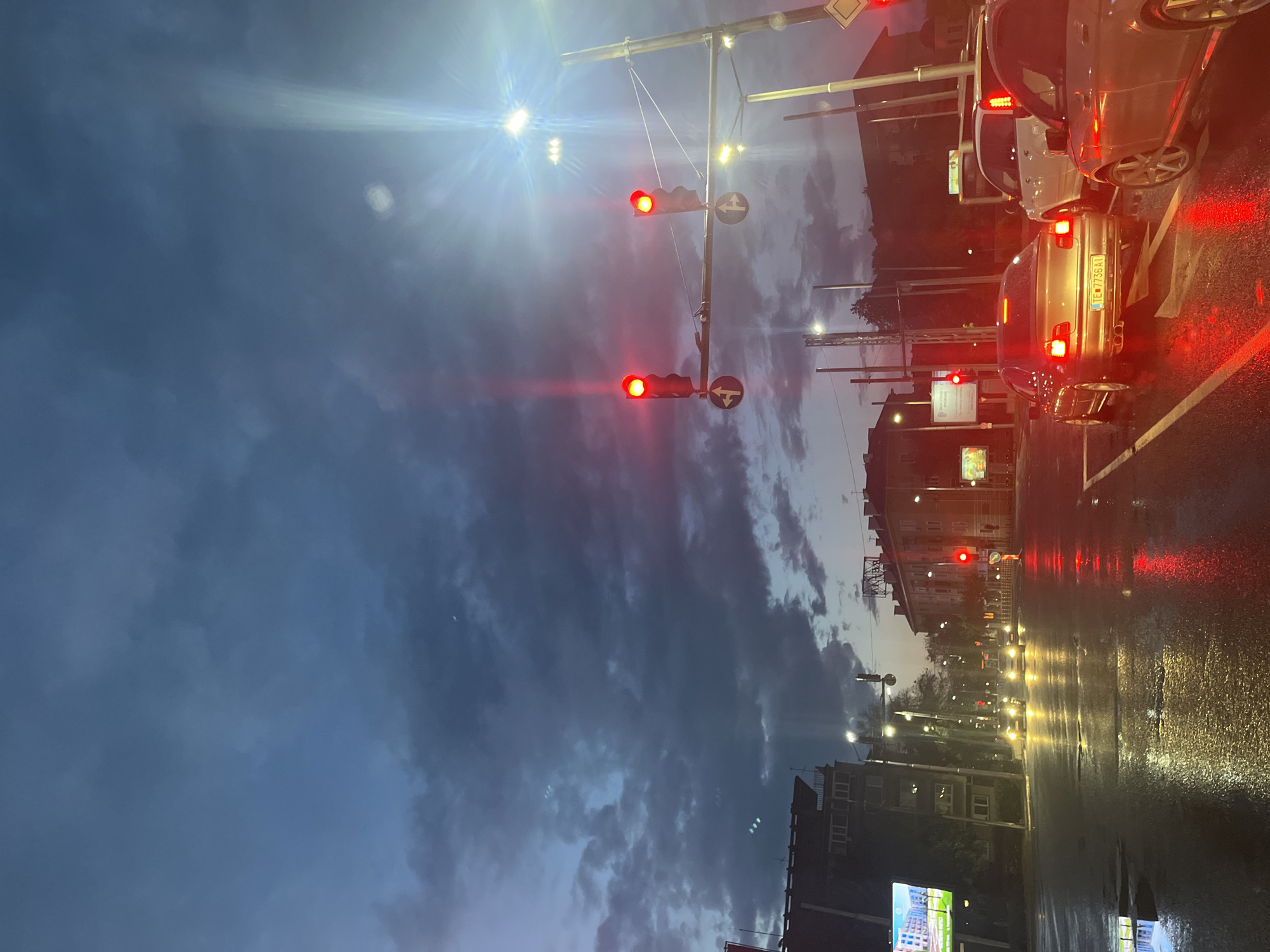 The image shows a street scene in Plovdiv, Bulgaria at dusk or dawn, after rain. The view is along a wet asphalt road, with cars stopped at a traffic light showing red. Reflections of lights are visible on the wet road surface. The sky is cloudy with a mix of dark and light patches. Buildings line the street on both sides, with streetlights and traffic signals visible. There are no people visible. The car license plate is "PB 738 KI".