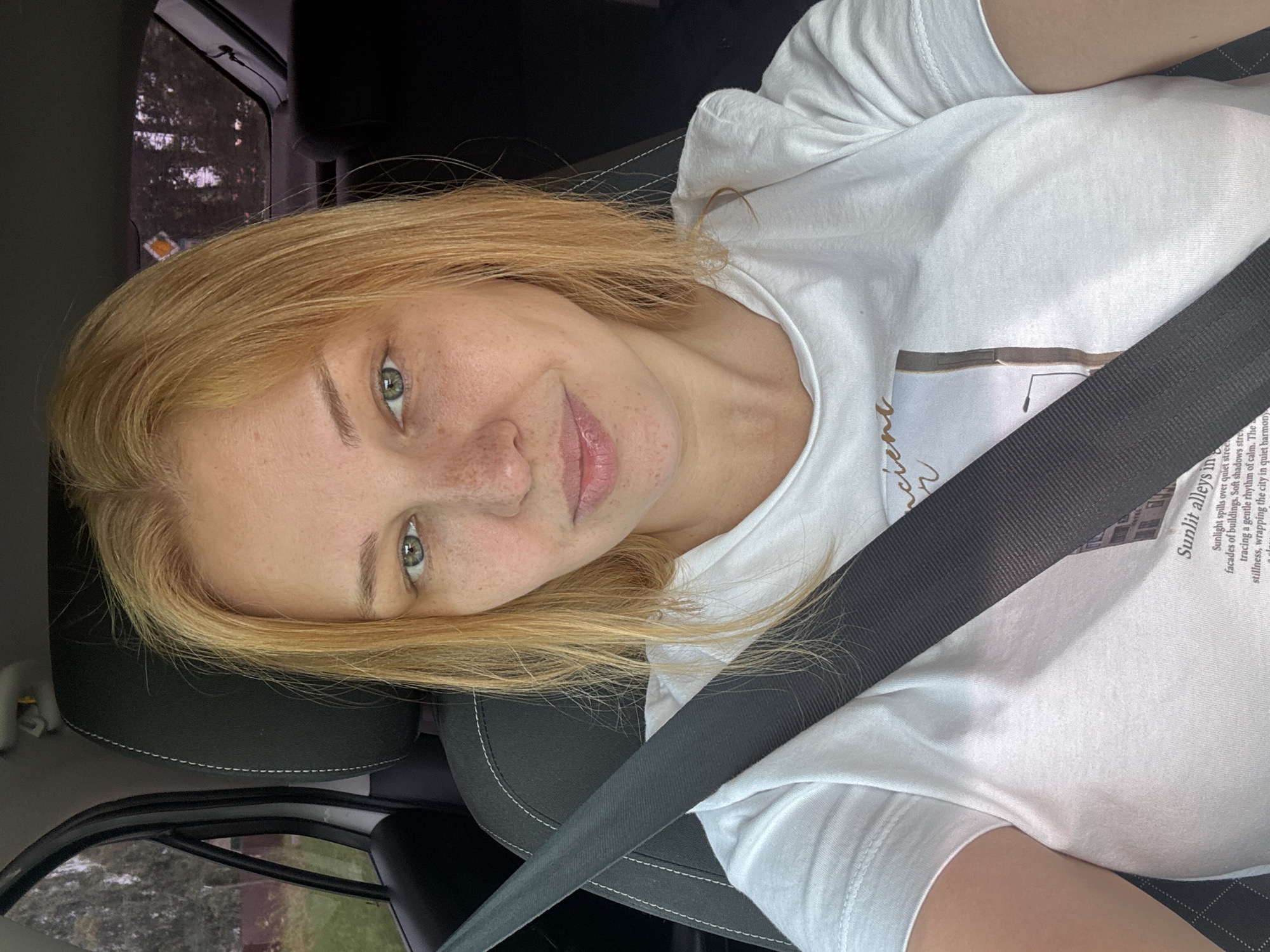 A close-up, slightly angled, medium shot captures a young Caucasian woman with blonde hair, fair skin with freckles, and green eyes, looking directly at the camera. She is seated in the driver's seat of a car, indicated by the presence of a black seatbelt crossing over her white t-shirt. The t-shirt has a printed design featuring text and an image of a building. The background is blurred, showing the interior of the car, including a black headrest and a partial view of the passenger side window with trees and foliage visible outside. The lighting suggests it is daytime. The image is an outdoor, daytime, candid photograph taken inside a vehicle. The specific location of Vereteya, Russia is not evident from the visual information provided in the image.
