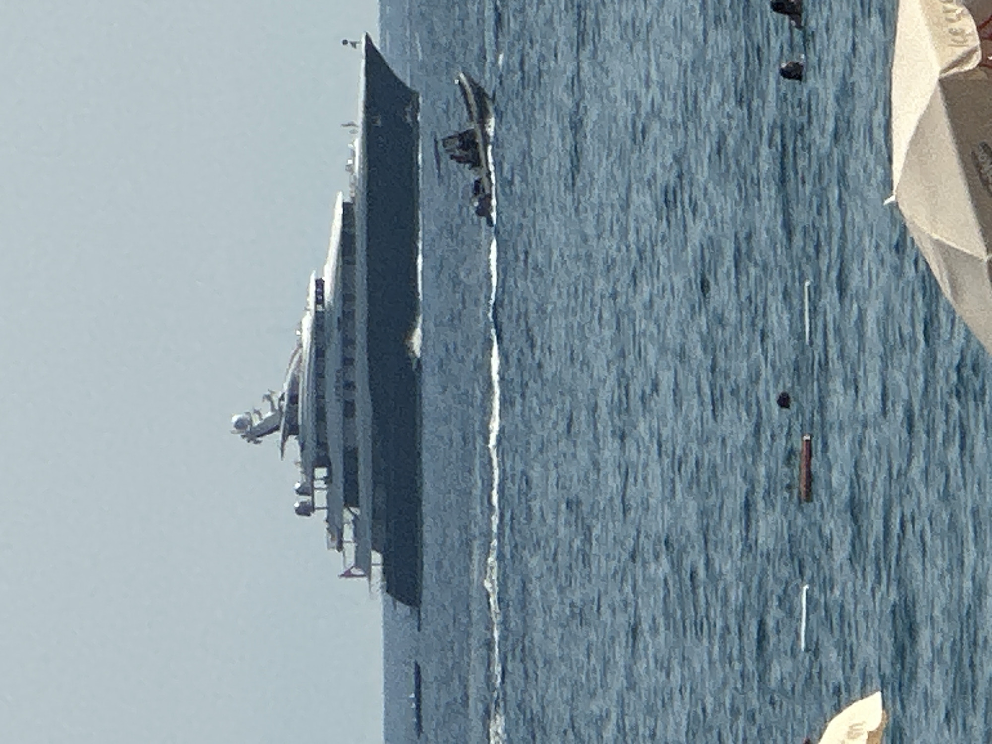 In the city of Boreti, Montenegro, a large dark-hulled yacht is anchored offshore on a clear day. The yacht features multiple decks and a radar dome on its upper level. In front of the yacht, a smaller, gray inflatable boat with a canopy and three individuals aboard is moving through the water, leaving a wake. Closer to shore, several buoys are visible in the blue water, along with a few swimmers. The foreground shows the top of a beige beach umbrella, indicating a shoreline presence. The overall scene depicts a tranquil marine environment with recreational activities.
