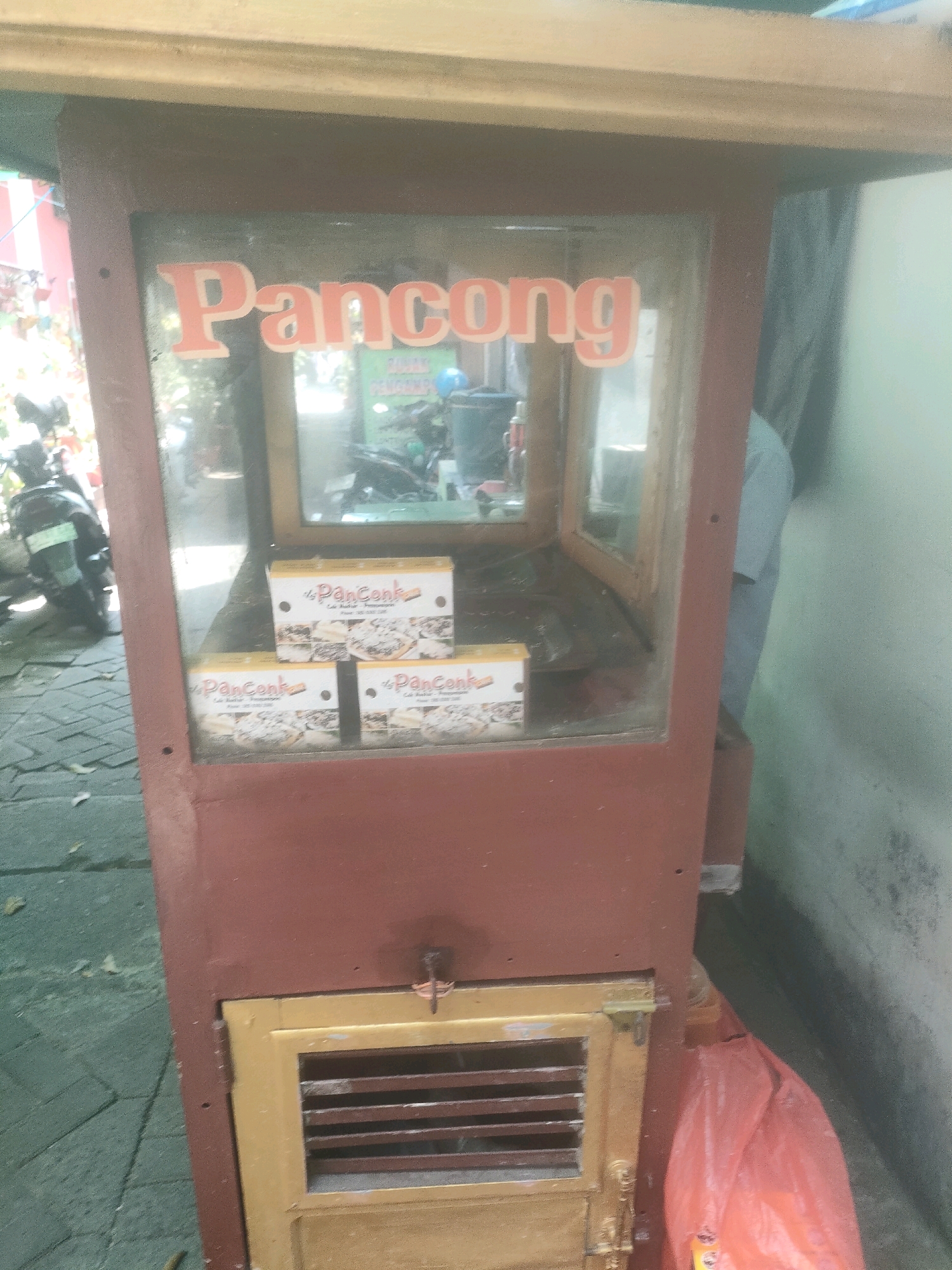 A red food cart with the word "Pancong" is displayed in front of a wall. The cart has glass windows where several boxes of "Pancong" are arranged. A red plastic bag is visible at the bottom right corner of the cart. The cart appears to be located on a cobblestone path outside, with greenery and a motorbike visible in the background. The lighting suggests it is daytime.