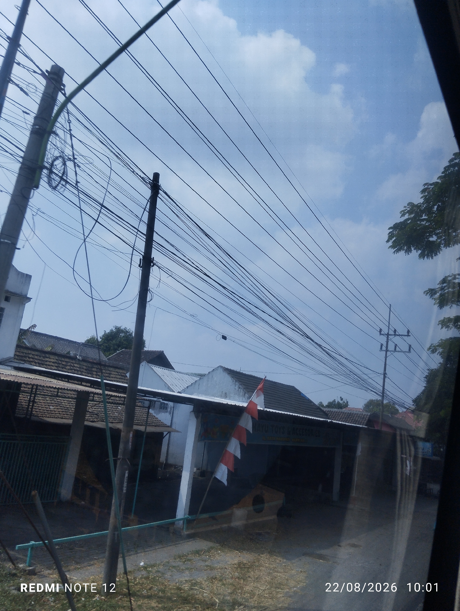 The image shows a street view in Bayemtaman, Indonesia. The foreground is dominated by a complex network of overhead power lines and utility wires, strung between poles and stretching across the frame. A concrete utility pole with attached wires is prominently positioned to the left of center, and another pole with insulators is visible further back and to the right.

In the middle ground, there are several buildings with tiled roofs. One building in the center has a white facade with a striped Indonesian flag (red and white) hanging on its side. A sign above the entrance to this building reads "JAYU TOYS & ACCESSORIES". The buildings appear to be shops or small businesses. A dark green metal fence is visible in front of some of these structures. The ground in front of the buildings is a mix of dry grass and dirt.

The background consists of a bright sky with scattered white clouds. Trees with green foliage are visible to the right of the frame.

The bottom left corner of the image displays the text "REDMI NOTE 12", and the bottom right corner shows a date and time stamp: "22/08/2026 10:01". There are no visible people in the image. The overall scene depicts an ordinary street in an Indonesian town, characterized by its infrastructure and commercial establishments.