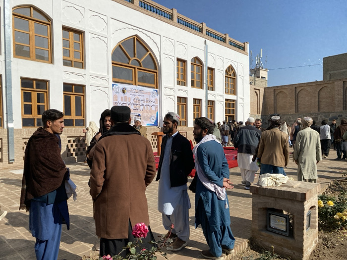 The image depicts an outdoor gathering of approximately 30-40 adult males within the **Ahmad Shahi Mosque Complex** in Kandahar City, Afghanistan. The scene unfolds under a clear blue sky on a bright day.
In the foreground, several men are engaged in conversation on a paved surface. To the left, a man wearing a brown shawl over a blue shalwar kameez faces right. Centrally, two men, one with a black cap and brown coat, the other with a dark head covering, have their backs to the viewer, conversing with a man to their right who wears a white turban-like head covering, a light blue shalwar kameez, and a white scarf.
To the left, a prominent two-story building features traditional Afghan architecture with a white facade, multiple arched and rectangular wooden-framed windows, and decorative upper molding. A large blue banner with white and yellow Pashto/Dari script and emblems is affixed to the building's front. To the right in the background, a traditional brick building with arched recesses is visible, behind which a brown structure topped with multiple communication antennas stands against the sky.
The ground consists of paved flagstones or bricks. In the lower right, a raised brick planter contains dark pink roses and some yellow flowers. This planter also features a small, framed, possibly illuminated sign and a piece of white cloth draped over its top. A red carpet is visible in the mid-ground, extending towards the background crowd, suggesting a designated path for an event. The men are predominantly dressed in traditional Afghan attire, including shalwar kameez, turbans, pakols, and various coats or shawls.