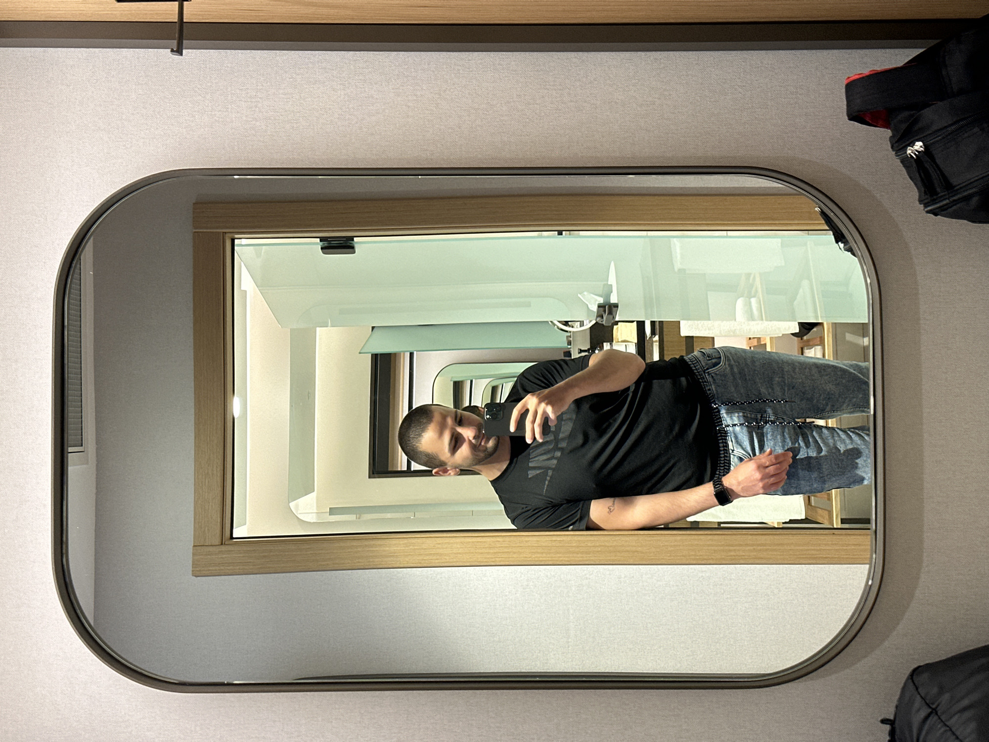 This image, captured in Razliv, Bulgaria, is rotated 90 degrees clockwise, depicting a man taking a mirror selfie. The primary subject is a man with a shaved head or very short dark hair and a beard, seen from the chest up. He is wearing a black t-shirt with a faint "NIKE" logo across the chest and blue jeans. A black smartwatch is visible on his left wrist. He holds a dark-colored smartphone to capture his reflection, showing a relaxed expression with a slight smile.
The man is reflected in a large, rounded rectangular mirror with a dark, thin frame. The mirror is mounted on a light-colored, possibly textured wall. Beyond his reflection in the mirror, an interior space is visible, featuring light-colored walls and wooden frames, suggesting a modern hotel room or apartment. There are also elements of glass or further reflections within this background.
In the original image's top right corner, a black bag, possibly a backpack, with a red lining is partially visible, suggesting it's placed near the mirror. A dark, slender bar or hanger is also seen at the very top edge of the image frame. The setting is indoors with ambient lighting.