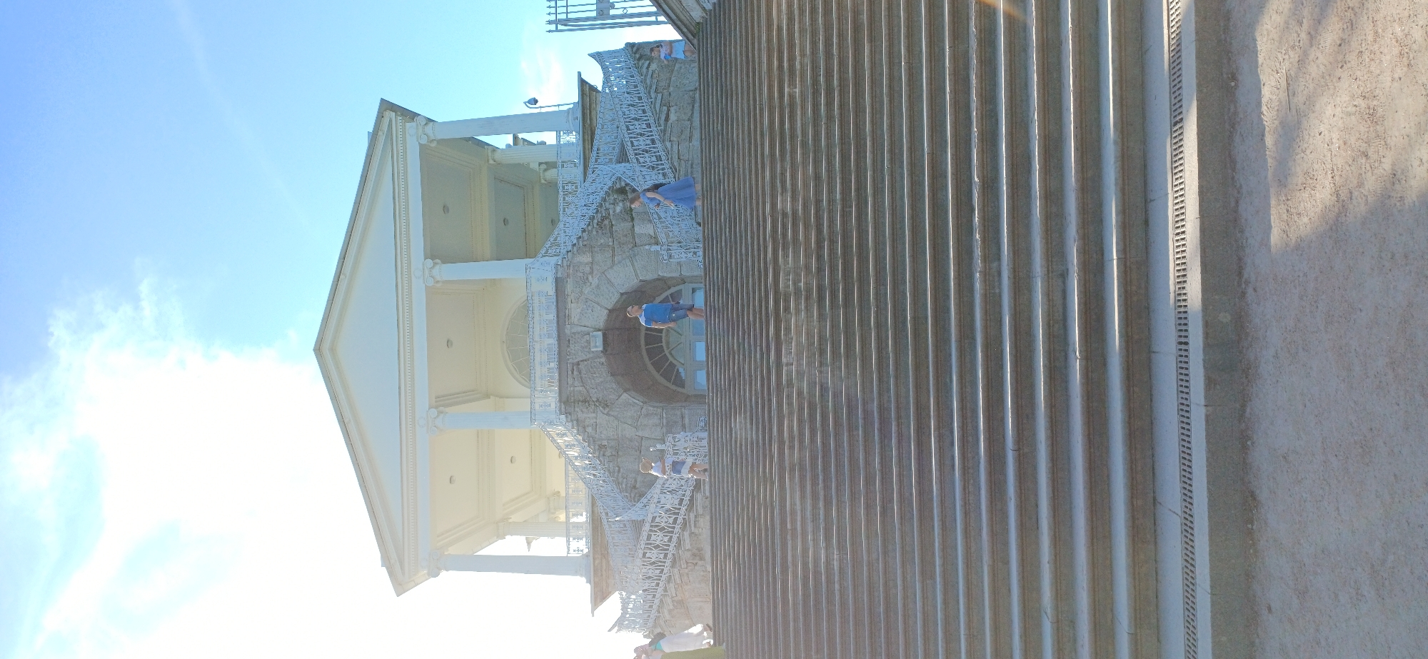 In Pushkin, Russia, a wide, grand stone staircase leads up to a neoclassical building with a colonnaded portico. The sun shines brightly, creating strong shadows on the steps. Several people are visible on the staircase and the upper level. On the left, a person in a white outfit is partially visible. Further up, a person in a blue and white outfit stands on the staircase. In the center of the upper level, a man in a blue t-shirt and shorts stands near a stone archway. To his right, a woman in a blue dress is walking up the staircase. On the far right, a person in a light blue garment stands on the upper level. The sky is a clear, bright blue with some wispy white clouds. The building appears to be a pavilion or a palace structure, consistent with the architectural style found in Pushkin's parks.