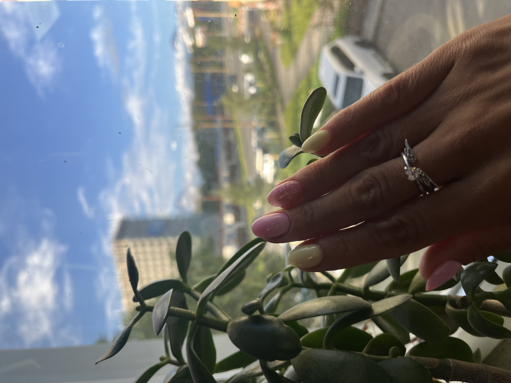 A close-up, low-angle shot shows a person's hand with manicured nails against a backdrop of a window overlooking a city. The hand, with nails painted in pastel pink, yellow, and pink glitter, is positioned in front of a jade plant. A silver ring with a small, clear stone and wing-like accents is worn on the ring finger. Through the window, a blurred cityscape is visible, featuring a tall building, trees, roads with cars, and a white SUV parked in the foreground. The sky is blue with scattered white clouds. The scene appears to be indoors, near a window, with the view suggesting an urban environment, possibly Petropavlovsk-Kamchatsky, Russia, given the context.
