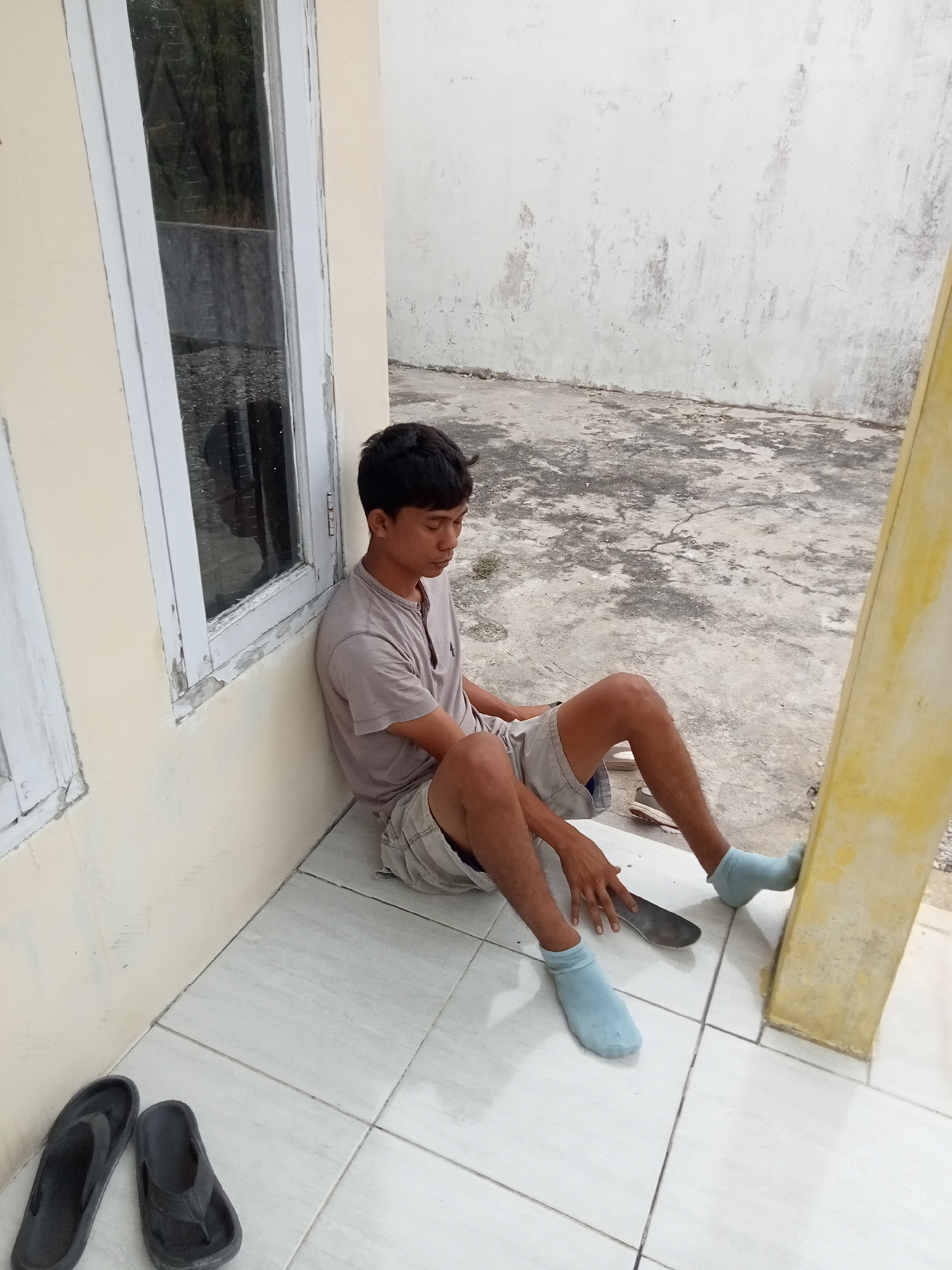 A young man of Southeast Asian descent sits slumped against a pale yellow wall, his head bowed and eyes closed. He is wearing a light brown t-shirt and grey shorts, with pale blue socks and dark flip-flops lying on the tiled floor nearby. In his right hand, he holds a large, flat metal object, possibly a spatula or a trowel. The man is positioned on a white-tiled patio, adjacent to a building with a weathered white window frame. The ground beyond the tiles is a rough, grey concrete surface. A weathered yellow pillar stands to the right of the man. The overall scene suggests a domestic or outdoor work area in Kualu, Indonesia, possibly a residential backyard or a small workshop.