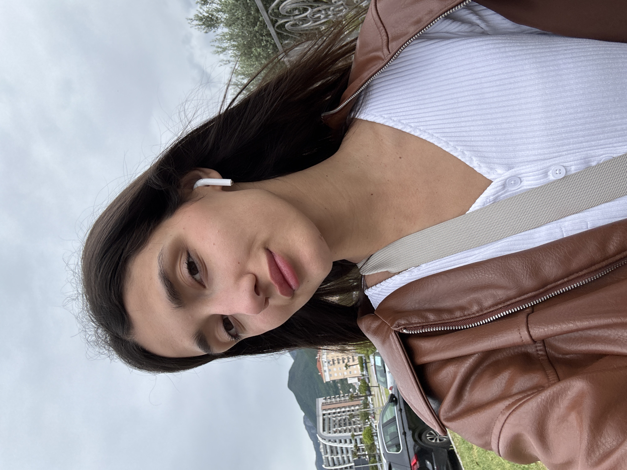 The image is a selfie of a young woman with long dark hair, wearing a white ribbed top, a brown leather jacket, and an earbud in her left ear. She is wearing a neutral-colored strap across her chest. The background shows a cityscape with buildings and a mountain in the distance, which identifies the location as Bar, Montenegro. A black van is visible behind her. The sky is overcast.
