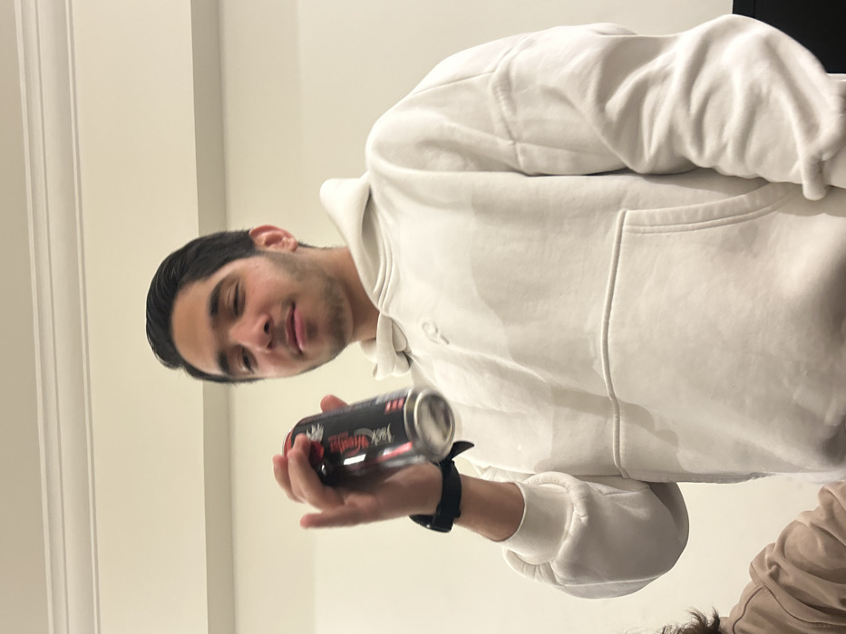 A young man in a white hoodie holds a black and red "Jack Daniel's" canned drink. He has dark hair and appears to be indoors. The background is a plain white wall and ceiling. Part of another person's shoulder and hair are visible in the lower right corner. He is also wearing a black wrist watch.