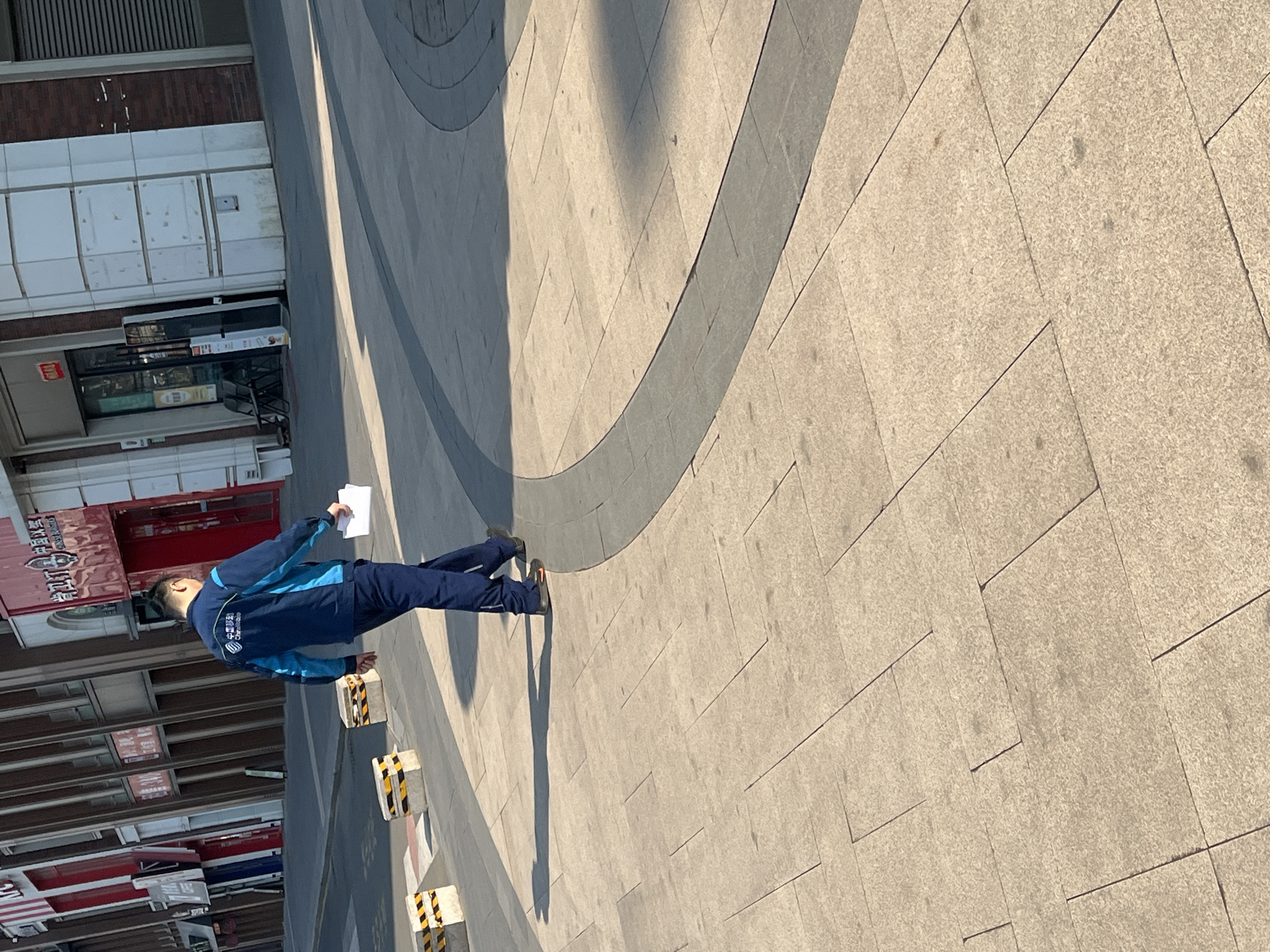 A single male individual walks from left to right across a light-colored paved plaza in Changchun City, China, under bright daylight. He is observed from behind, wearing a dark blue and light blue uniform jacket with "中国移动" (China Mobile) branding on the back, dark blue trousers, and dark shoes with orange sole accents. He carries a stack of white papers in his right hand. The paved surface beneath him consists of large, light-tan rectangular and square tiles, featuring a distinct curved dark gray paved strip. A prominent shadow of the individual is cast to his left. In the midground, three black and yellow striped concrete barrier blocks are positioned near a dark asphalt road. The background reveals a row of multi-story commercial buildings, including a KFC restaurant identifiable by its red and white logo on the far left, and other storefronts displaying Chinese characters, with facades ranging from red to brown and white. Two dark chairs are visible outside a building entrance on the right.