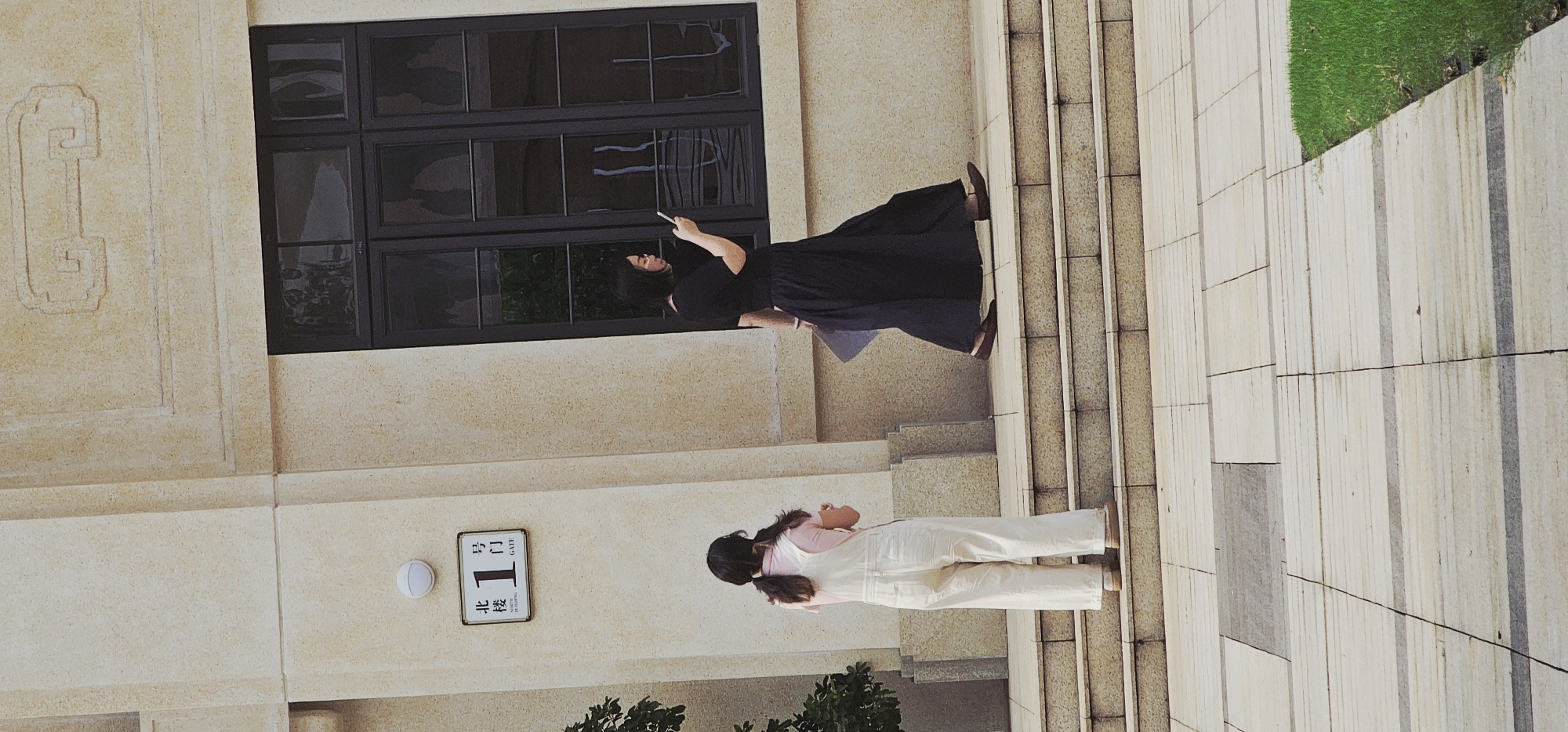 The photograph depicts two individuals on or near a staircase adjacent to a building in Yuzhong District, China. The building has a light-colored façade with dark-framed windows and a decorative relief. A sign with Chinese characters and the number "1" is visible on the building. One individual, dressed in a long black dress, appears to be using a mobile phone. The other person is wearing overalls and a pink top. The location includes a staircase leading to a grassy area.