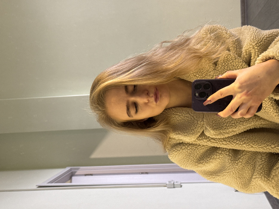 An adult female is depicted in a mirror selfie, positioned indoors. She has long, light blonde hair parted to her right, falling over her shoulders. Her eyes are closed or cast downwards, and her lips are slightly pursed. She is wearing a thick, light beige or cream-colored textured coat, possibly a fleece or sherpa material. Her right hand holds a dark-colored smartphone, likely an iPhone Pro model with a visible triple-camera array, positioned in front of her mid-section. The phone case appears dark purple or black with faint white text "PARIS" visible on the lower left of the case. Her fingernails are painted a dark reddish-brown hue. The background reveals a light greenish-grey wall, with a partial view of an open white doorframe and door on the left side of the frame. A dark floor, possibly grey or black, is visible at the bottom left. The location is an indoor space, likely a private residence or hotel room, in the city of Unknown, Unknown.
