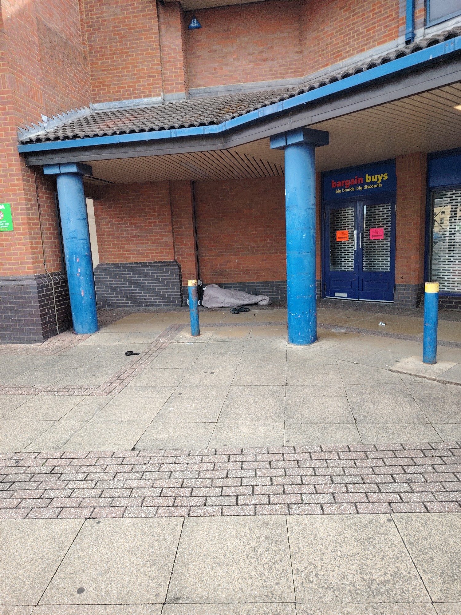 The image depicts the entrance area of a commercial building in Peterborough, United Kingdom. The foreground features grey concrete paving slabs with a central strip of reddish-brown brick pavers. A single black flip-flop or sandal lies on its side on the grey paving in the mid-ground.
The building itself is constructed from reddish-brown brickwork, with a darker grey brick base. A curved canopy, featuring dark roof tiles and a blue gutter, extends over the pedestrian area. Below the canopy is a white corrugated soffit. Anti-bird spikes are installed along the left edge of the roofline. Two large, weathered blue cylindrical pillars support the canopy; one is integrated into the building on the left, and a narrower one stands further right. A smaller blue bollard with a yellow top is positioned to the right of the freestanding pillar. A blue "Chubb" security sign is affixed high on the brick wall to the right.
To the right, a dark blue double door, currently secured by closed metal shutters or grates visible through glass, serves as the entrance to a "Bargain Buys" store. The store's sign, in red, yellow, and orange text, reads "big brands, big discounts" in white. Orange and pink notices are taped to the inside of the door.
Under the canopy, between the two blue pillars and to the left of the store entrance, a person is visible lying on the ground, covered by a grey blanket or sleeping bag. A pair of black sandals are placed beside them. Small pieces of litter are scattered on the paved area near the store entrance. A green sign is partially visible on the far-left brick wall.