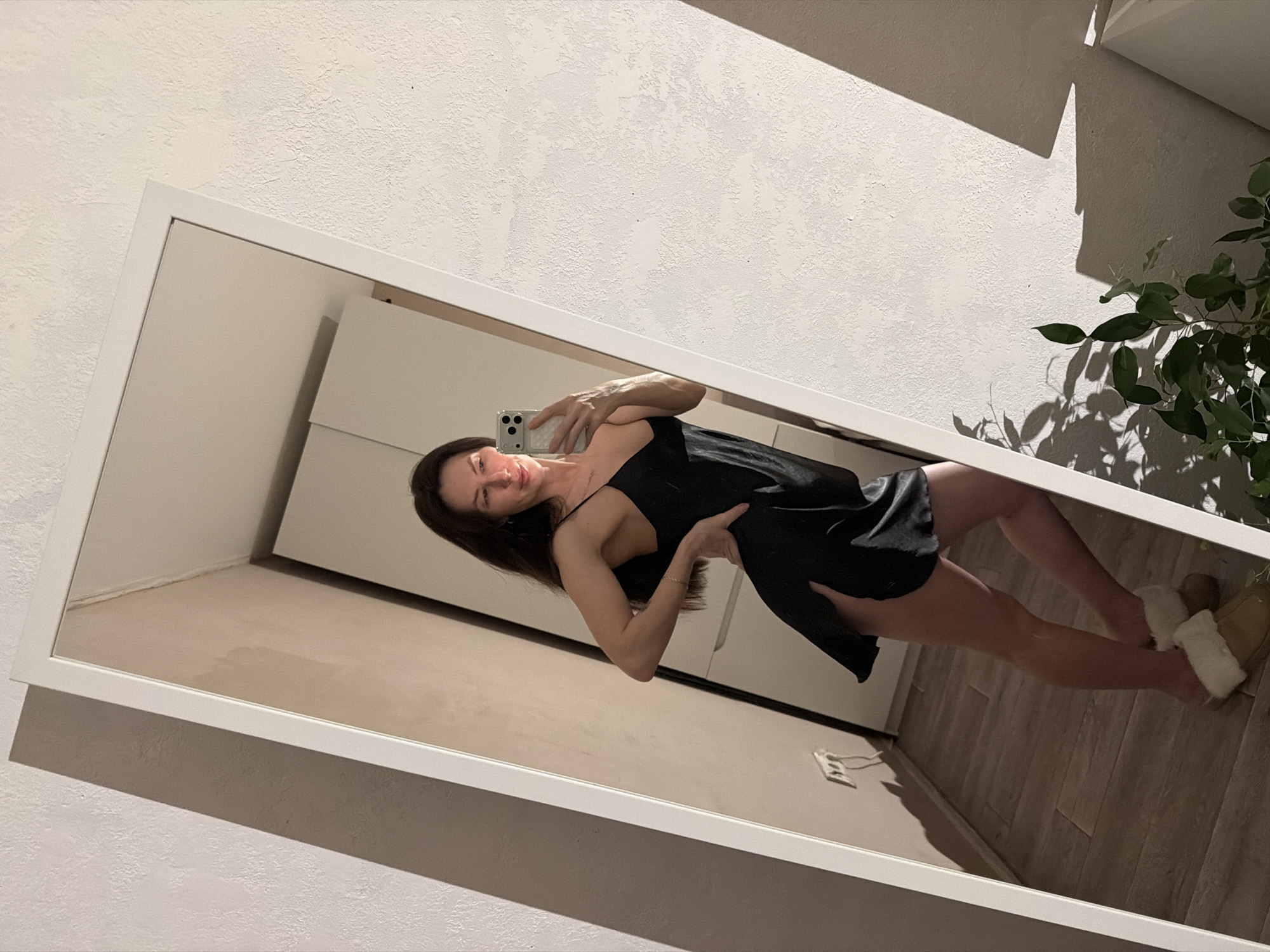 A woman stands in front of a mirror, holding a smartphone and taking a selfie. She is wearing a short black slip dress and slippers. The background includes a white wall, a closed white cabinet, a section of wood flooring, an electrical outlet, and a potted plant. The reflection in the mirror reveals the room setting. The city of Perm, Russia is not discernible from the image.