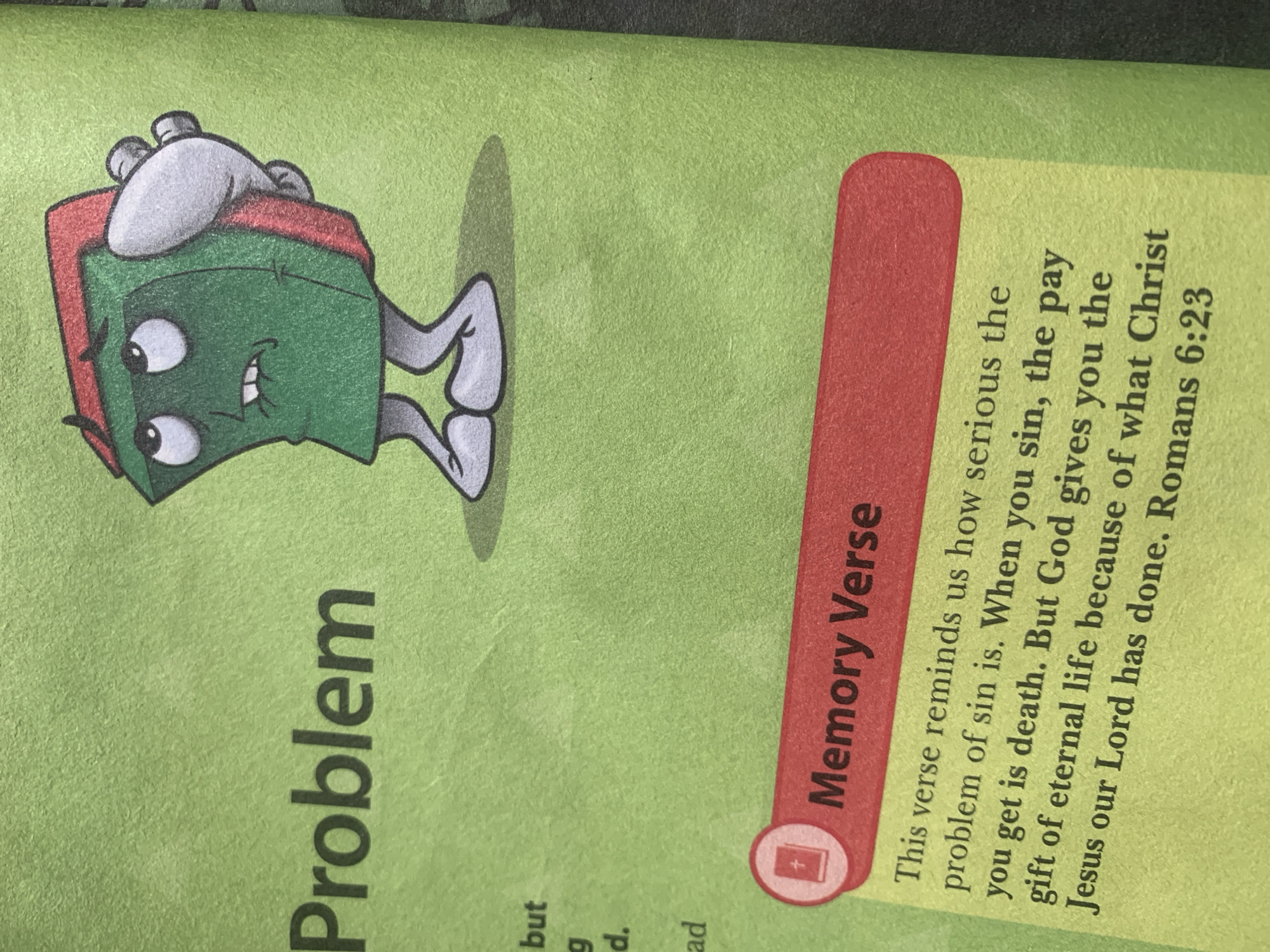 The image shows a page from a children's book or tract with a green background. The top portion of the page features a cartoon illustration of a green book with red edges, a distressed expression, and arms behind its back, standing on two legs. To the left of the illustration, the word "Problem" is printed in large, dark letters. Below "Problem," a few lines of text are partially visible, including "but," "g," "d.," and "ad."
The bottom portion of the page contains a section titled "Memory Verse" highlighted in a red banner with a stylized book icon and a cross. Below this banner, a passage of text is printed in black. The visible text reads: "This verse reminds us how serious the problem of sin is. When you sin, the pay you get is death. But God gives you the gift of eternal life because of what Christ Jesus our Lord has done. Romans 6:23."
The image does not contain any information about the city of Jauro Yinu, Nigeria, or any people present. The scene is static and contained within the depicted page.