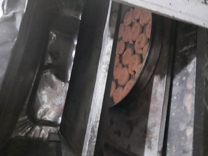 A glimpse inside an industrial oven reveals metal racks and foil lining. A circular tray, positioned on one of the racks, holds numerous round, light orange food items, possibly baked goods. The interior of the oven appears dimly lit and utilitarian. Metal surfaces show signs of wear and use.