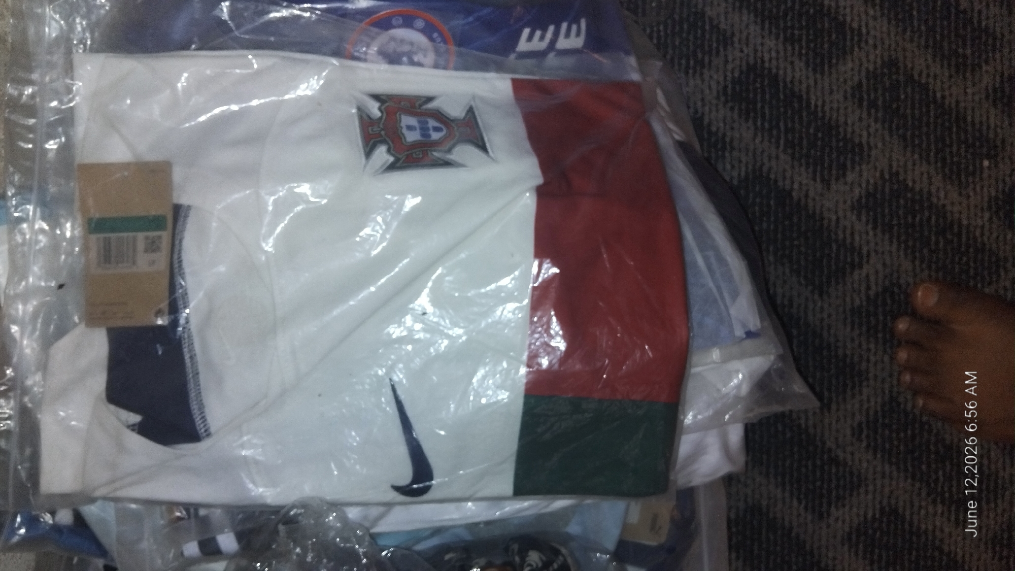 A white t-shirt with a dark blue collar is visible through plastic packaging. The t-shirt features a dark blue Nike logo on the left chest and the Portuguese coat of arms on the right chest. The lower portion of the t-shirt has a horizontal stripe that is divided into a dark green section on the left and a red section on the right. The t-shirt is laid flat on a dark, textured surface. A timestamp in the bottom left corner of the image reads "June 12, 2026 6:56 AM." A bare foot is partially visible in the bottom right corner of the image. The scene appears to be indoors, possibly in a storage area or a retail setting, though the specific location is unknown.