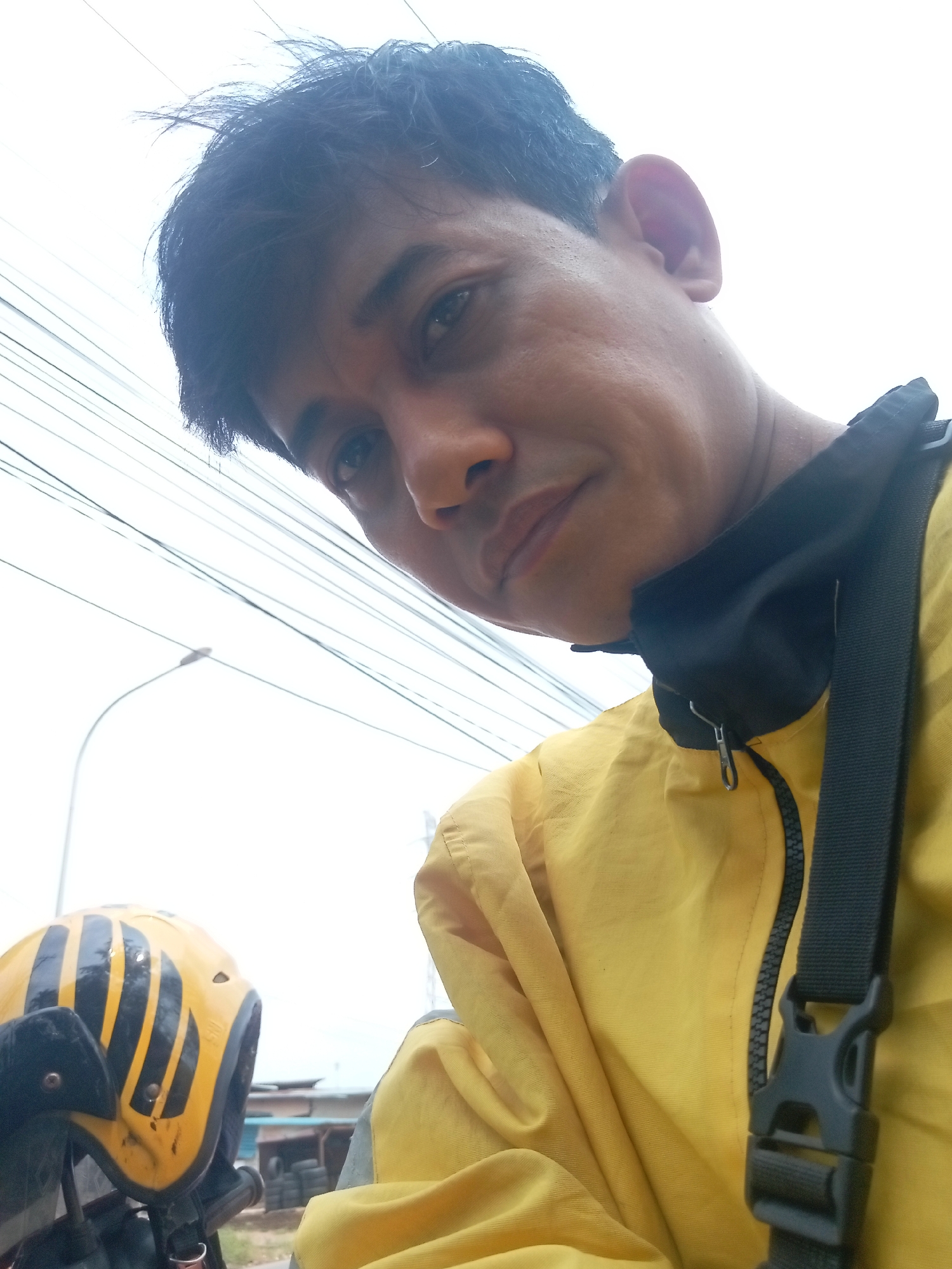 A close-up, low-angle selfie captures a man with short, dark hair and brown eyes wearing a bright yellow jacket with a black collar. He has a dark strap with a black buckle crossing over his chest. The man's face is partially obscured by the angle and his clothing. In the lower left of the frame, a yellow and black striped helmet is visible, suggesting the man may be on a motorcycle. The background is a bright, overcast sky with numerous overhead wires crisscrossing the frame. A faint silhouette of a street lamp can be seen in the upper left quadrant. The overall impression is of an outdoor scene, likely in an urban environment given the wires and lamp post, possibly during the daytime. The location is identified as Batam, Indonesia.