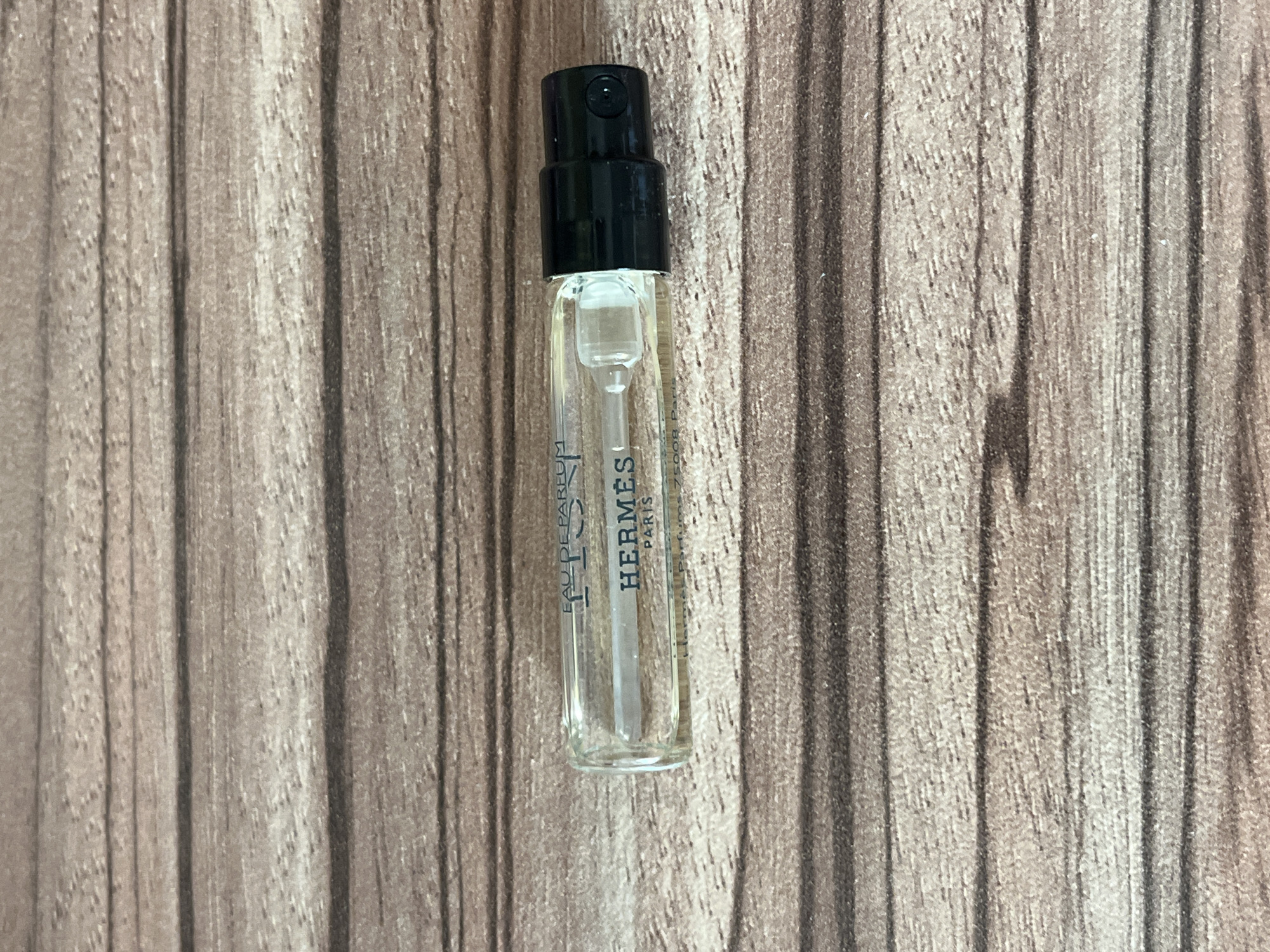 A small, clear glass perfume sample vial with a black plastic sprayer top is centered on a light brown wooden surface. The wood features a distinct vertical grain pattern and a textured, slightly worn appearance. The vial contains a clear liquid, and a thin, clear dip tube extends from the sprayer mechanism into the liquid.
Prominently displayed vertically on the side of the vial is the brand name "HERMÈS" in black capital letters, with "PARIS" directly beneath it. Above "HERMÈS," the text "EAU DE PARFUM" is partially visible. Further text below "PARIS" is largely obscured or illegible, but fragments suggest a street address like "75008 Paris" and product details such as "Natural Perfume".
The image is a straightforward product shot, focusing on the perfume sample. No people are present, and no activity is depicted. The lighting is even and consistent, highlighting the vial's contents and the background texture. The overall aesthetic is clean and functional. The location context for this media file is Sofia, Bulgaria.