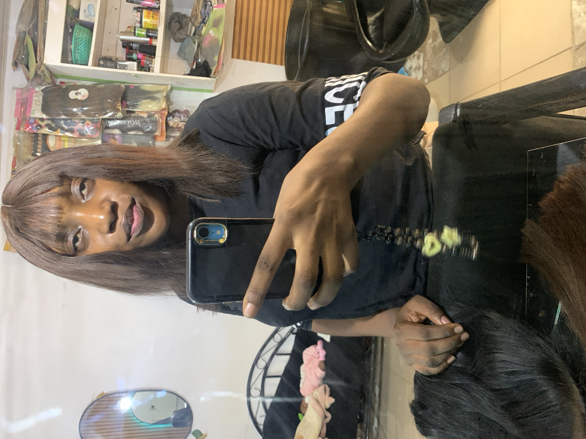 The image shows a person with medium-brown hair and dark skin taking a selfie in what appears to be a hair salon. The person is wearing a black t-shirt and is holding a black smartphone with a gold rim. In the background, there is a shelf with various hair products and wigs. A hair dryer chair and a stylist chair are visible. The reflection of the mirror shows a glimpse of what appears to be the interior of the salon. Based on the information provided, it's not possible to confirm this location is indeed the city of Jos, Nigeria.