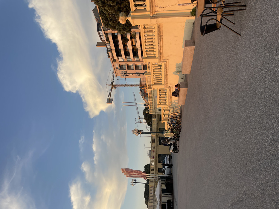 This image depicts an outdoor public area, likely a promenade, in Aldoar, Foz do Douro e Nevogilde, Portugal, under a clear blue sky with scattered, elongated white clouds, illuminated by warm, low-angle sunlight originating from the left. The ground surface is dark asphalt. Partial views of dark outdoor cafe chairs and a table are visible on the extreme right.
In the midground, a light-yellow concrete retaining wall with a decorative balustrade extends horizontally, exhibiting visible cracks in its surface. Two light-colored, rectangular concrete benches are positioned along this wall. One individual is observed lying prone on the left bench, partially covered by dark fabric and a light brown object. Another person is seated on the ground adjacent to the right bench, partially obscured. To the left of the benches, several bicycles are parked in a rack, alongside a white scooter. One person in dark attire stands near the scooter. Further left, a white car is parked, with two individuals standing in its vicinity. A white tented outdoor seating area with tables and chairs is visible on the far left. Multiple ornate, multi-globular lampposts and three tall poles displaying red banners are positioned behind the vehicles and structures.
The background reveals multi-story residential or commercial buildings situated on an elevated terrain. Two prominent yellow tower cranes are visible against the sky, positioned behind the buildings, indicating ongoing construction. Green foliage from trees is present on the hillside to the right of the buildings.
