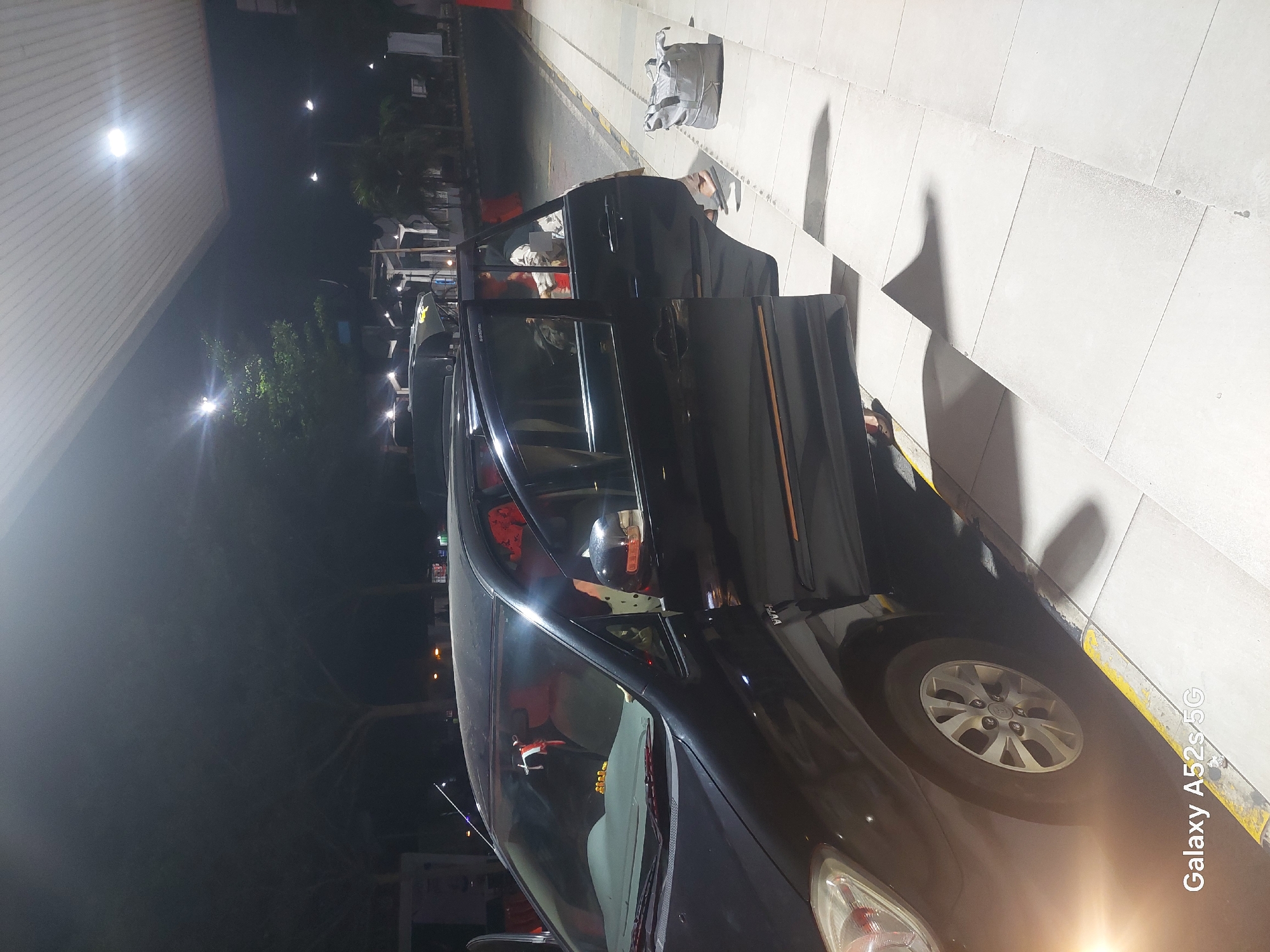 In Bumisari, Indonesia, at night, a black car is parked on a paved surface, possibly a gas station or parking lot, under an overhang with lighting. The car's front driver's side door is open, and a person is seen exiting or entering the vehicle. Another person is visible in the driver's seat. A grey duffel bag rests on the ground near the open door. The scene is illuminated by artificial lights.