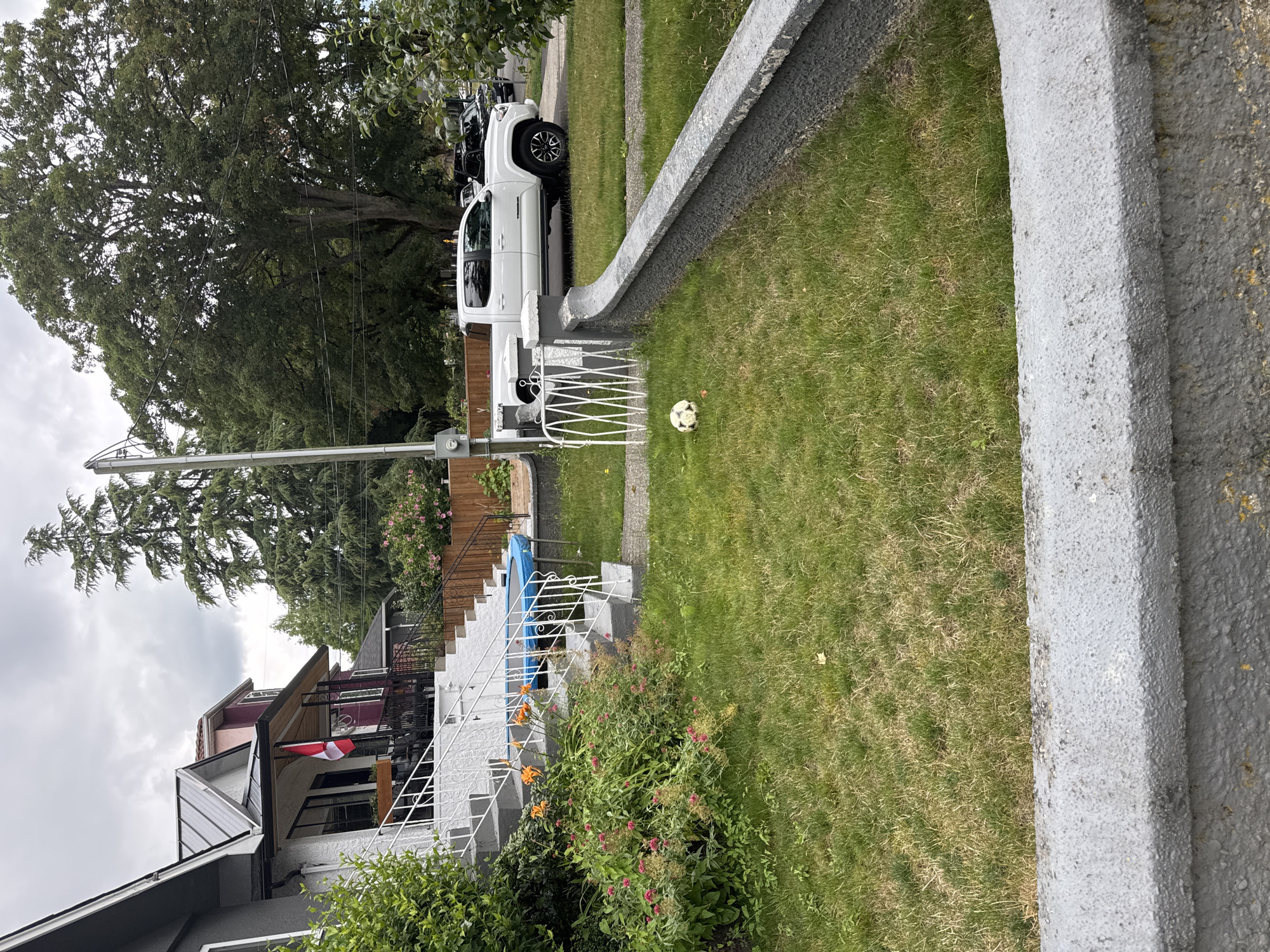 A white pickup truck is parked in the driveway of a residential property in Vancouver, Canada. The truck is facing towards the right of the image. In the foreground, a soccer ball lies on the grass in front of a low, grey, textured wall. Behind the truck, other vehicles are parked on the street. The house has a white facade and a grey metal roof. A Canadian flag is displayed on the porch of the house. Green trees and bushes are visible in the background. The sky is overcast.