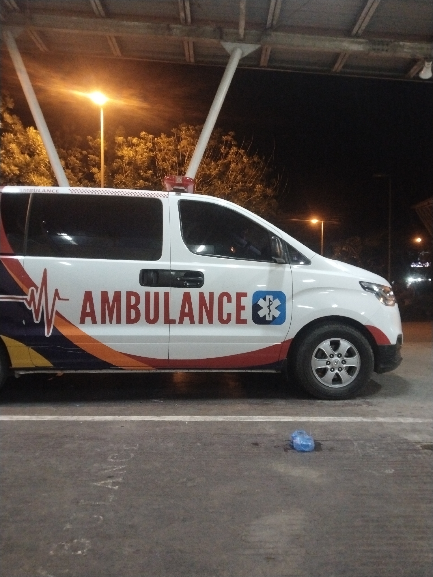 An ambulance with the word "AMBULANCE" printed in large red letters on its side is parked under a shelter in Banda Aceh, Indonesia. The background includes night sky, trees, and street lights. A small blue plastic bag lies on the ground in front of the ambulance.